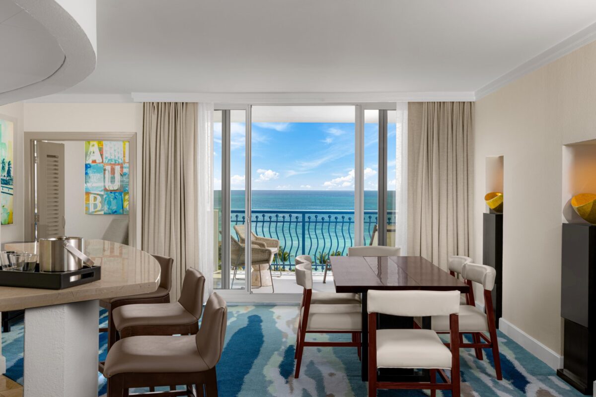 A modern hotel suite with a dining table, chairs, and a balcony overlooking the ocean. Large windows let in natural light, and the room features contemporary decor with abstract art and light-colored furniture.