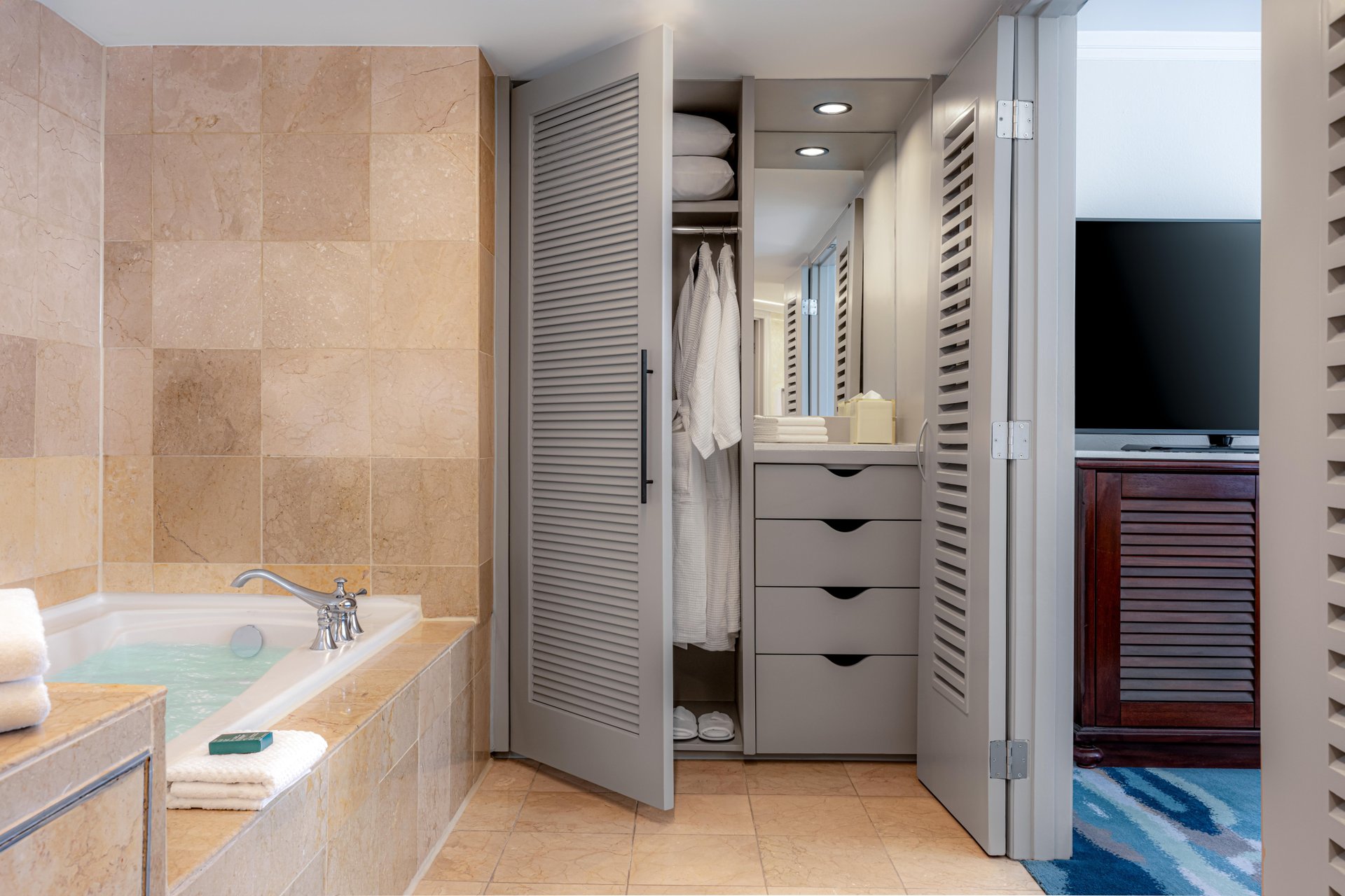 A modern bathroom with beige tile, a bathtub filled with water and towels on the edge, and an open closet showing robes, shelves, and neatly folded towels. A TV and dresser are visible in the adjoining room.
