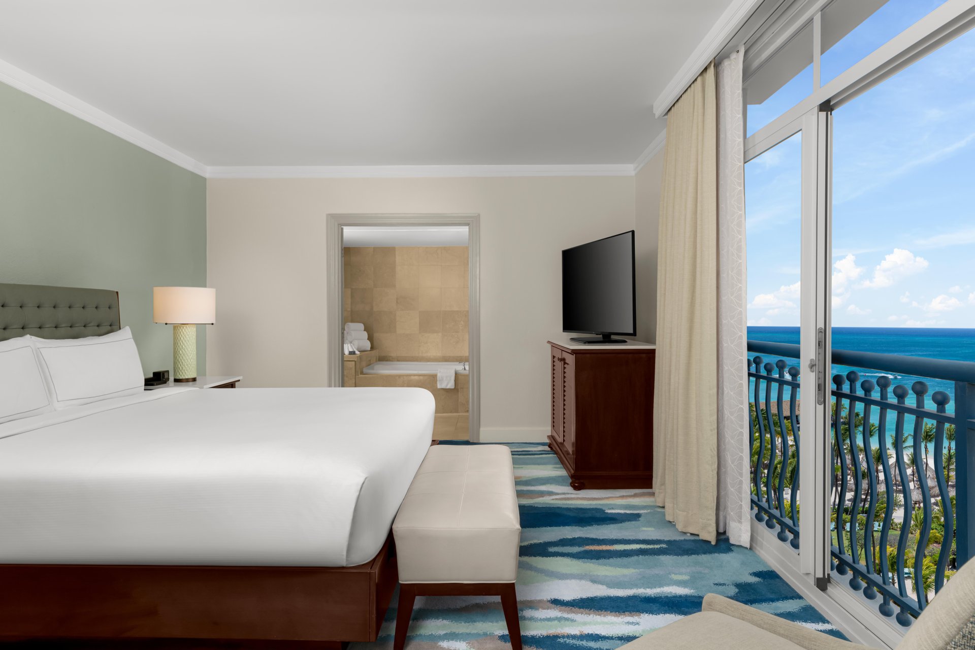A hotel room with a large bed, upholstered bench, flat-screen TV, and a view of the ocean through floor-to-ceiling windows; an open doorway reveals a bathroom with a soaking tub.
