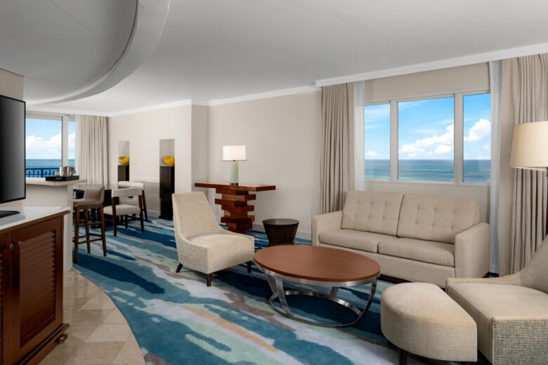 A modern hotel suite with a beige sofa, armchairs, and a wooden coffee table, features large windows overlooking the ocean and a blue-patterned carpet, with a dining area visible in the background.