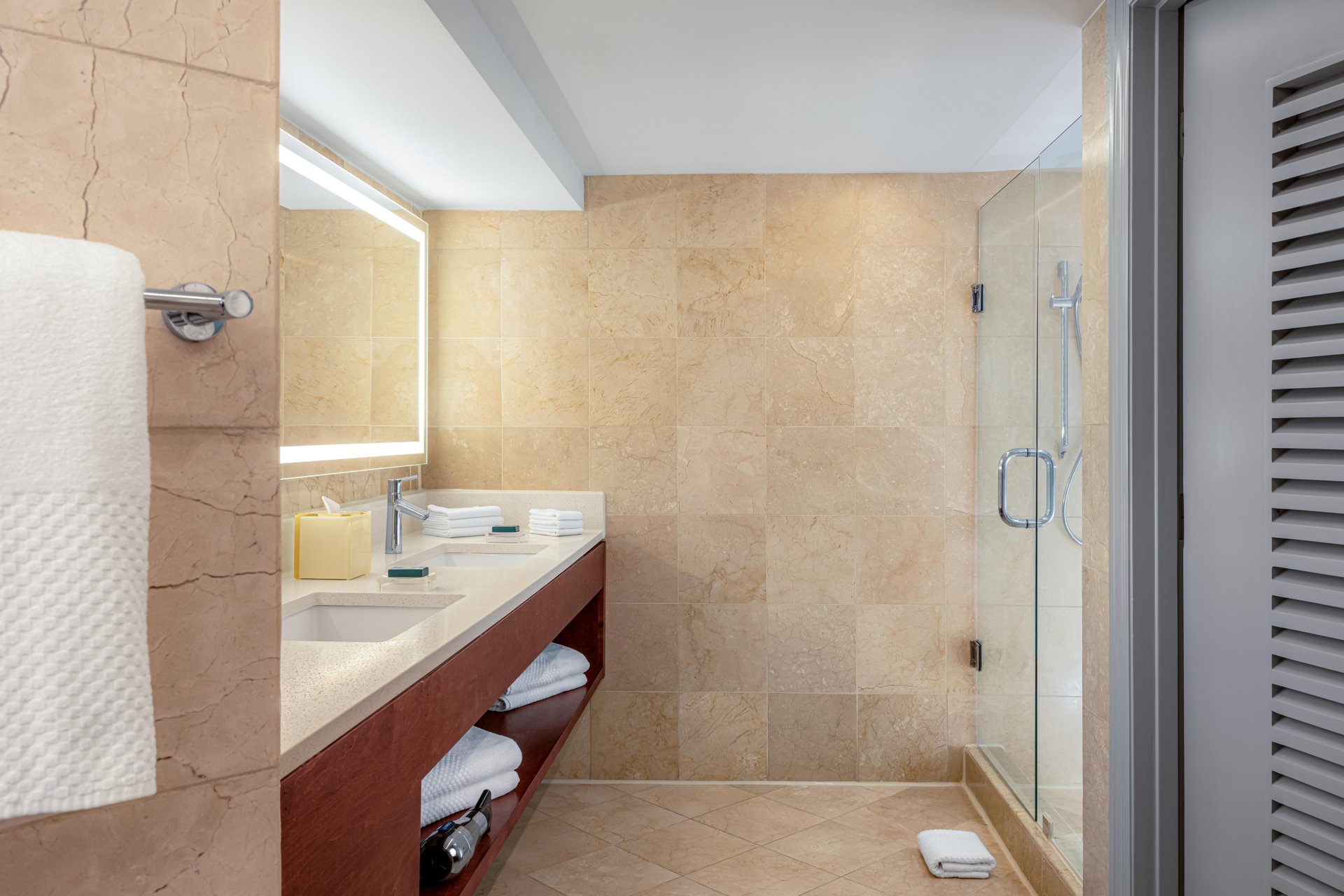 Modern bathroom with beige tile walls, a double sink vanity with towels and toiletries, a large illuminated mirror, and a glass-enclosed walk-in shower. A towel hangs on the wall next to the vanity.