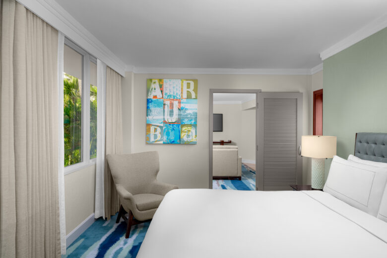 A modern hotel room with a large bed, armchair by a window, abstract art on the wall, bedside lamp, and an open door leading to a sitting area. Light colors and a blue-patterned carpet decorate the space.