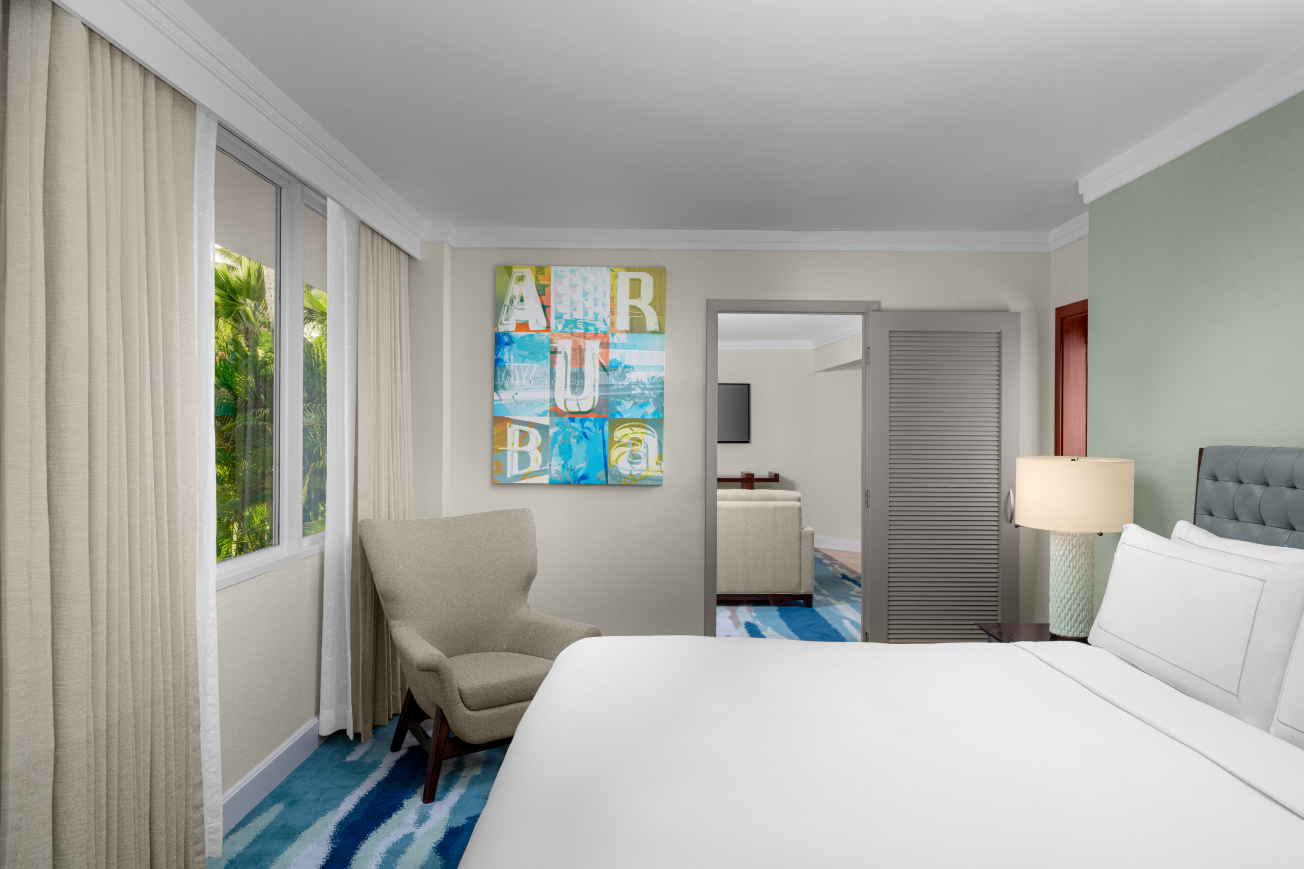 A modern hotel room with a large bed, armchair by a window, abstract art on the wall, bedside lamp, and an open door leading to a sitting area. Light colors and a blue-patterned carpet decorate the space.