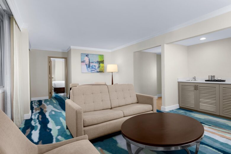 A modern hotel suite featuring a beige sofa, armchair, and round coffee table on a blue-patterned carpet, with a wet bar and bedroom visible in the background. Art hangs on a cream-colored wall.