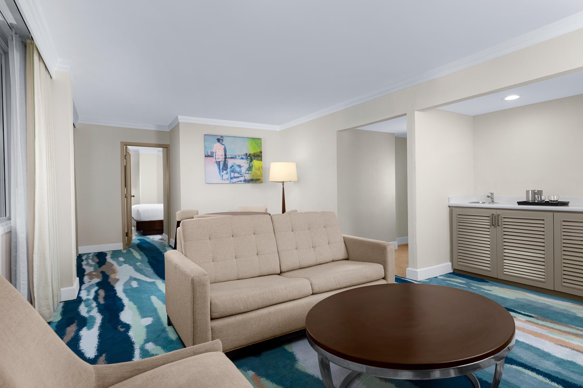 A modern hotel suite featuring a beige sofa, armchair, and round coffee table on a blue-patterned carpet, with a wet bar and bedroom visible in the background. Art hangs on a cream-colored wall.