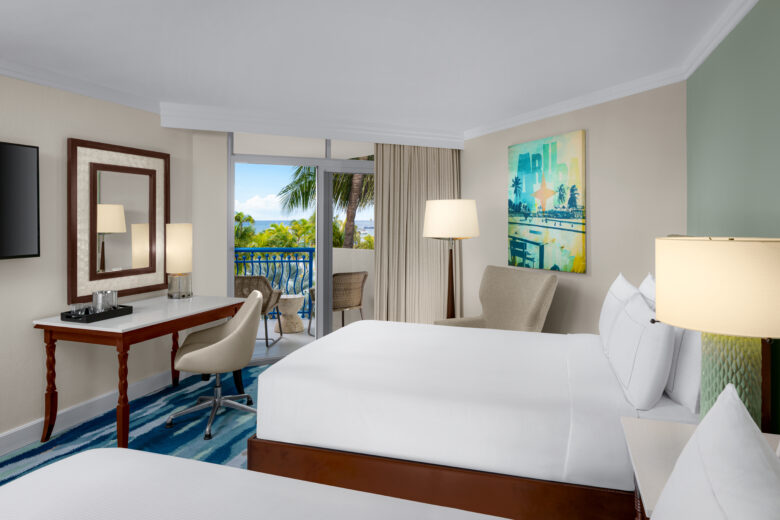 Modern hotel room with two double beds, a desk with a chair, a flat-screen TV, armchair, lamps, colorful wall art, and a balcony with a view of palm trees and a blue sky outside.