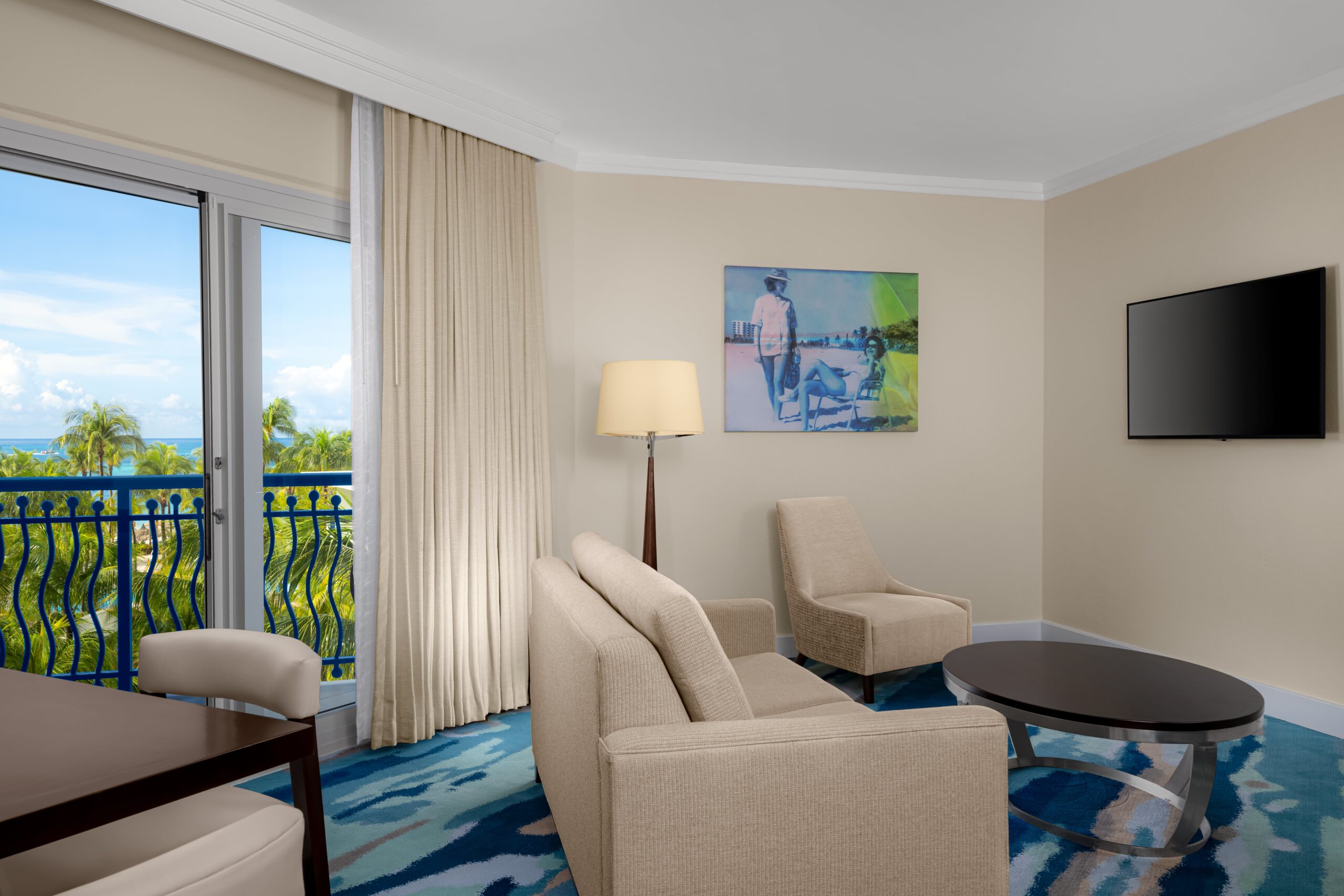 A bright hotel room with two beige armchairs, a sofa, a coffee table, a wall-mounted TV, a lamp, a desk, and large windows with sheer curtains overlooking palm trees and blue sky. Artwork hangs on the wall.