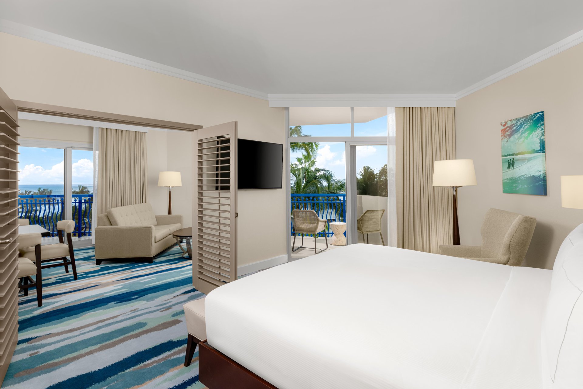 A bright hotel room with a king bed, a sitting area with chairs and a sofa, a wall-mounted TV, modern decor, large windows, and a balcony overlooking palm trees. Blue, white, and beige tones dominate the space.