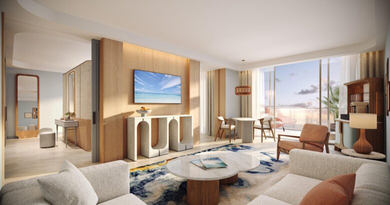 A modern, sunlit living room with neutral tones, wooden accents, a wall-mounted TV, a round coffee table, a patterned rug, and large windows overlooking a balcony with ocean views.