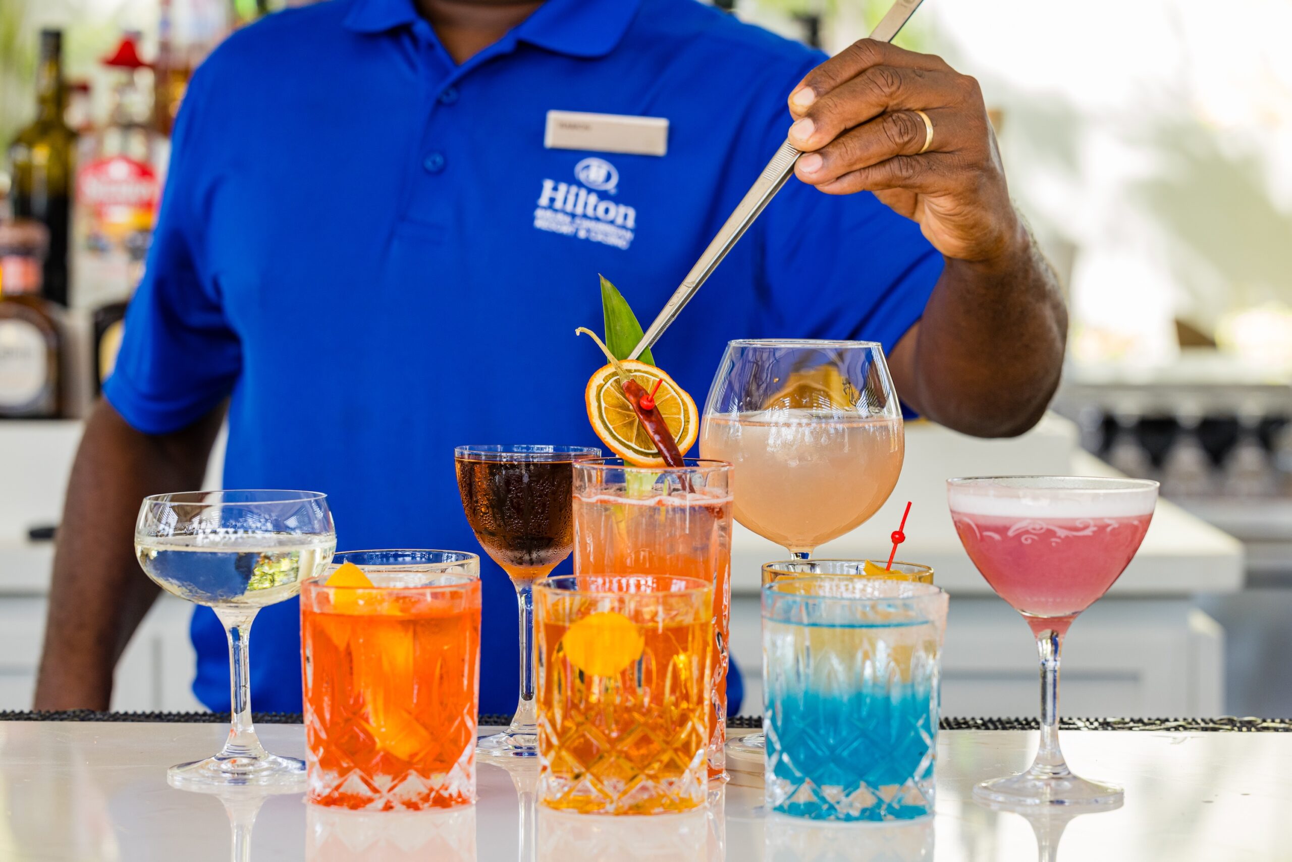 A bartender in a blue Hilton shirt decorates a colorful array of cocktails and drinks on a bar counter, using tongs to place a garnish on a large glass. Bottles are visible in the background.