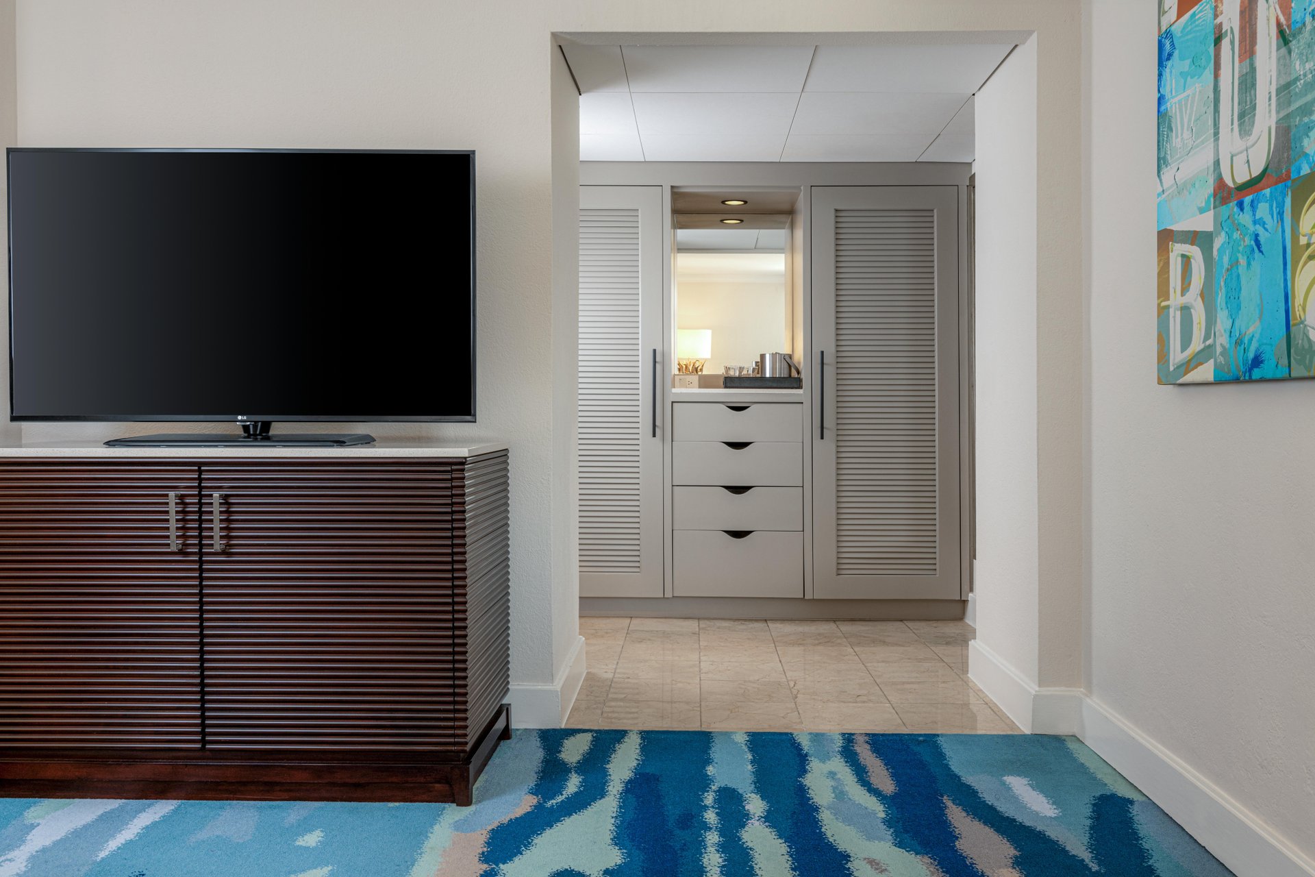 A hotel room with a large flat-screen TV on a wooden cabinet, a blue patterned carpet, and a view into a small kitchenette area with white cabinets and louvered doors. A colorful abstract painting hangs on the wall.