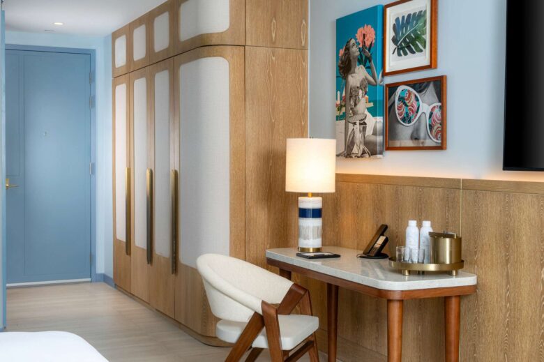 A hotel room features a wooden wardrobe, a small desk with a chair, a lamp, water bottles, and a tray. Artworks and photos hang on the wall above the desk.