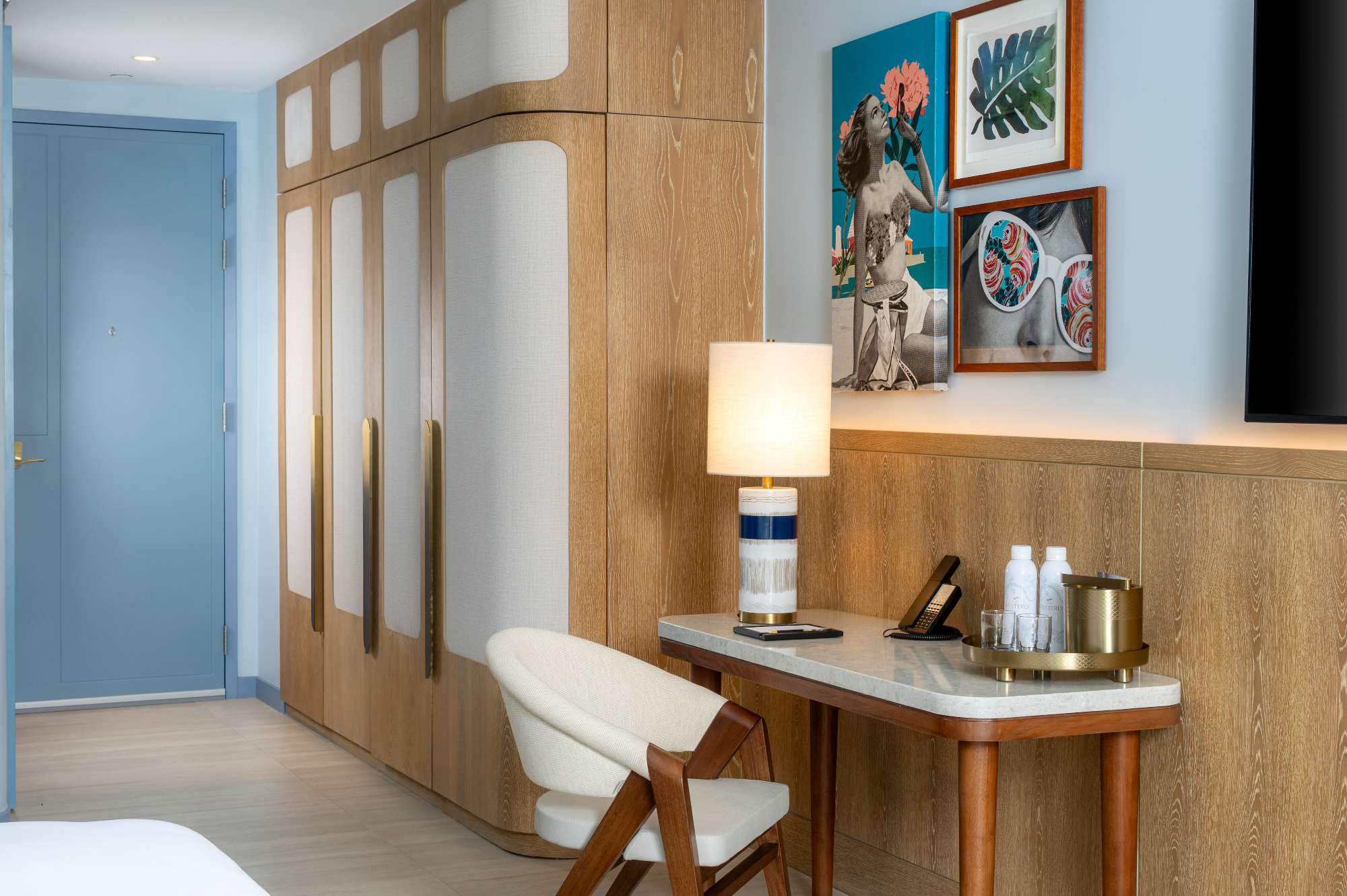 A hotel room features a wooden wardrobe, a small desk with a chair, a lamp, water bottles, and a tray. Artworks and photos hang on the wall above the desk.