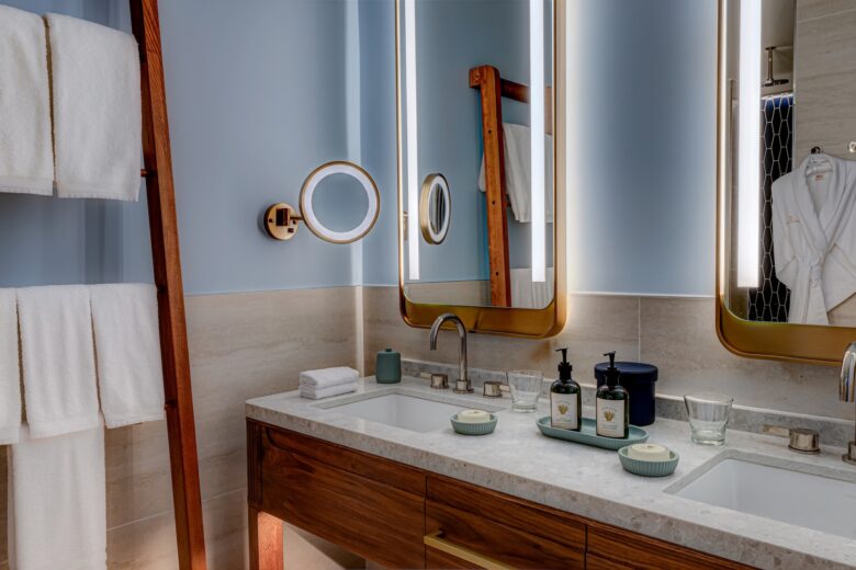 Modern bathroom with a double sink vanity, large mirrors with lights, towels hanging on a wooden rack, two soap dispensers, glass cups, and a robe hanging on the wall in the background.