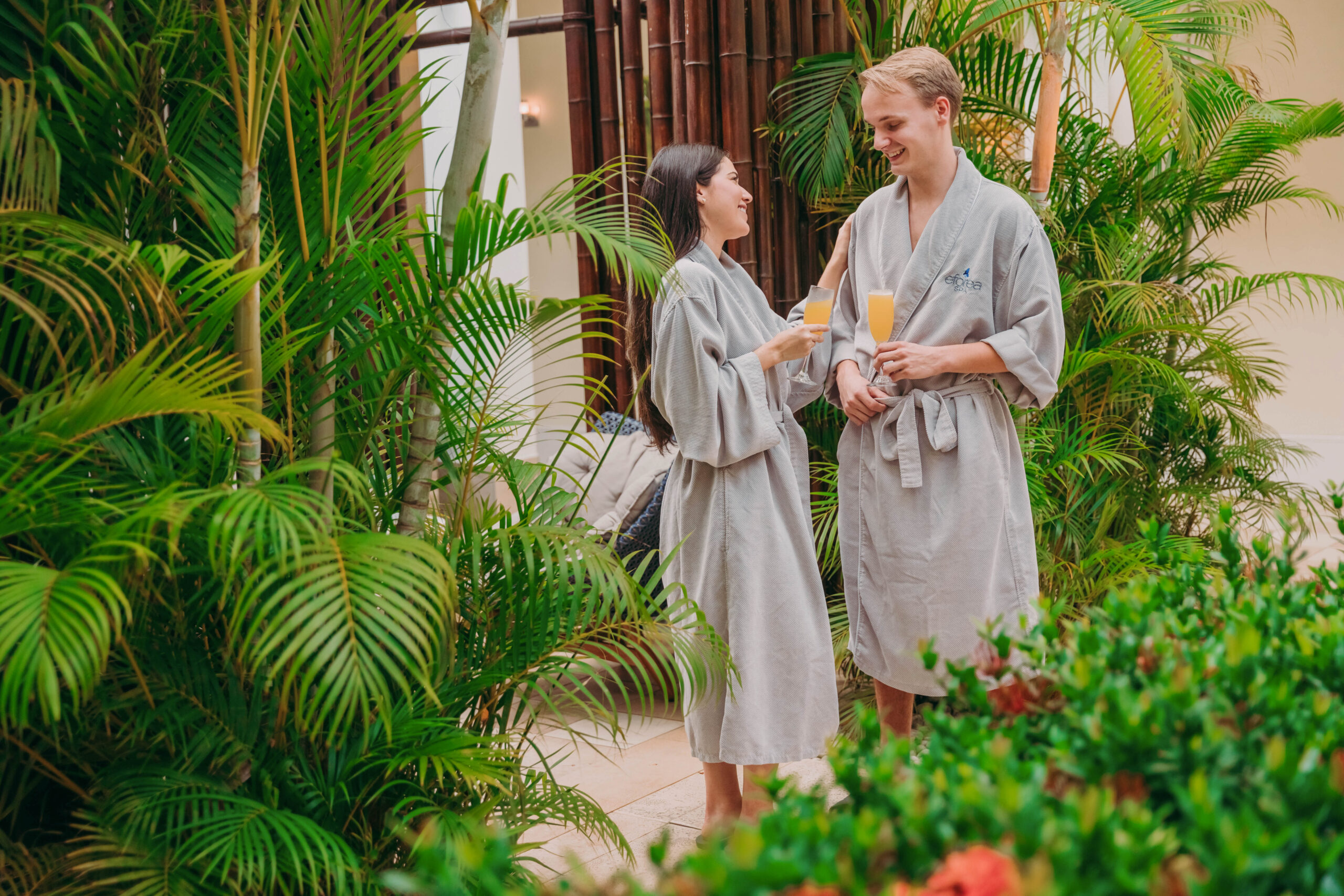 A man and woman in gray robes stand among lush green plants, smiling and holding glasses of orange drinks, appearing relaxed and happy in a tropical spa or garden setting.