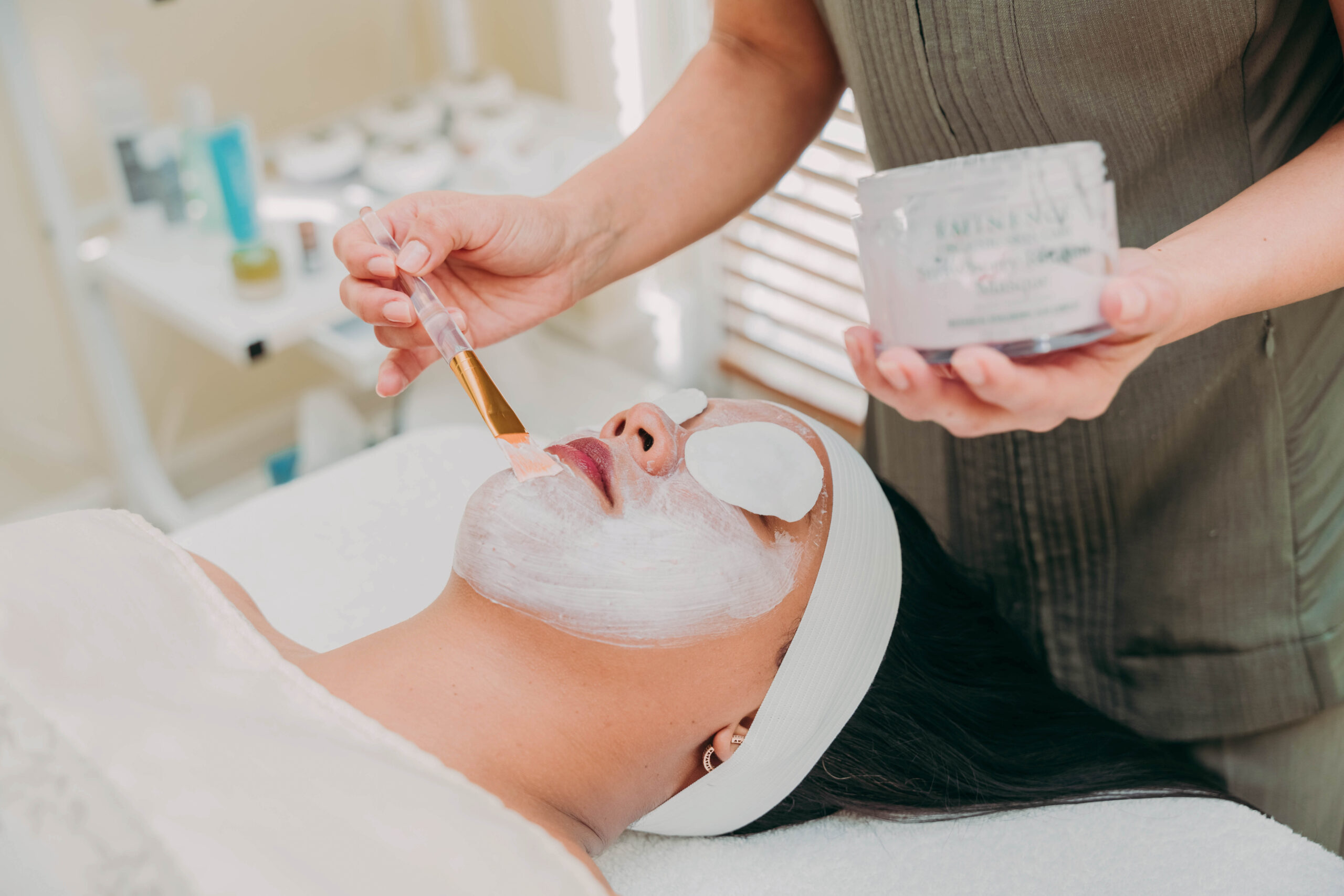A person with a headband and eye pads lies on a spa bed while a facial mask is applied with a brush by an esthetician holding a jar of skincare product. The setting appears clean and calming.