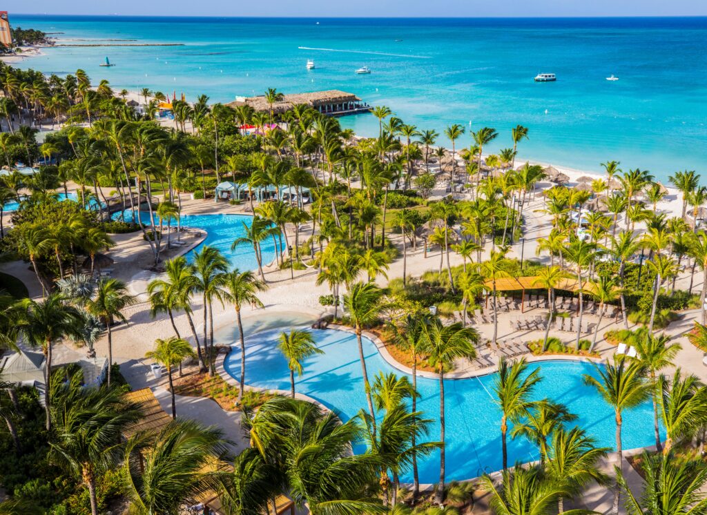 Aerial view of a tropical beach resort with palm trees, swimming pools, cabanas, and loungers by the turquoise ocean, with boats visible on the water and clear blue skies above.