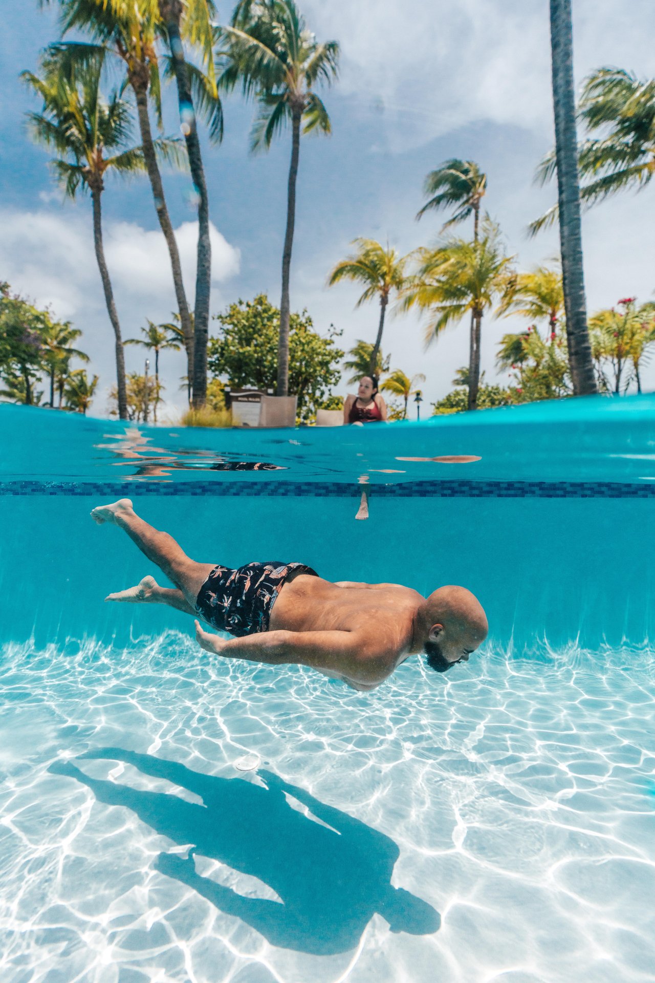 A man in swim trunks swims underwater in a clear pool, with sunlight creating patterns on the pool floor. Palm trees, blue sky, and a child standing at the pool edge are visible above the water.