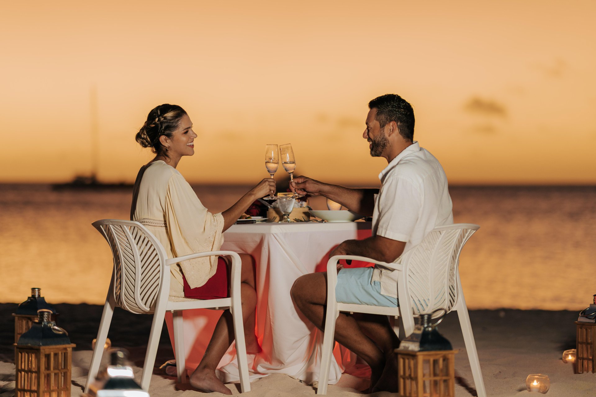 A couple sits at a table on the beach, toasting with wine glasses at sunset. The ocean and a sailboat are in the background, with lanterns placed around their table.