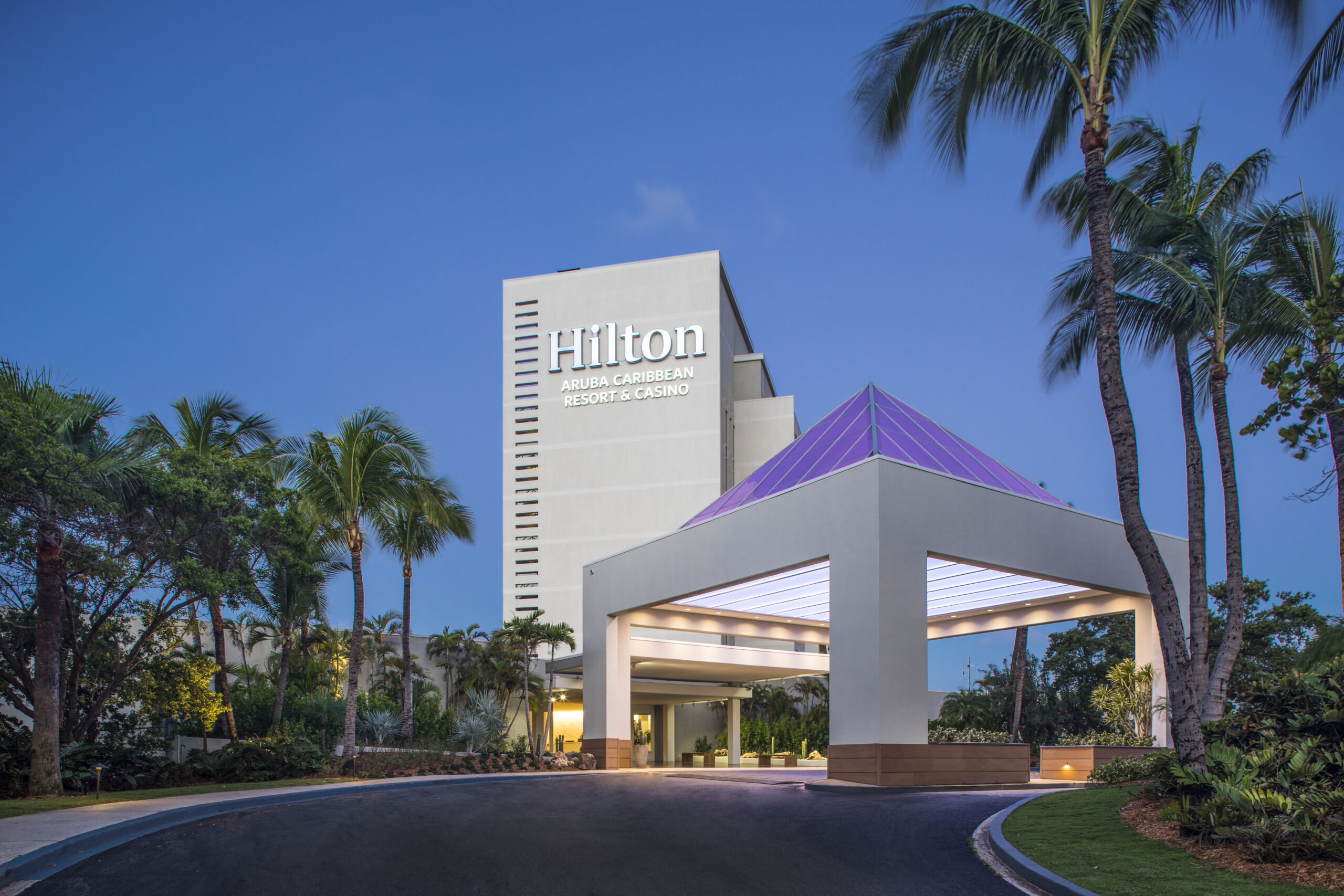 The entrance of the Hilton Aruba Caribbean Resort & Casino at dusk, with a modern white building, a covered driveway, and palm trees against a clear blue sky.
