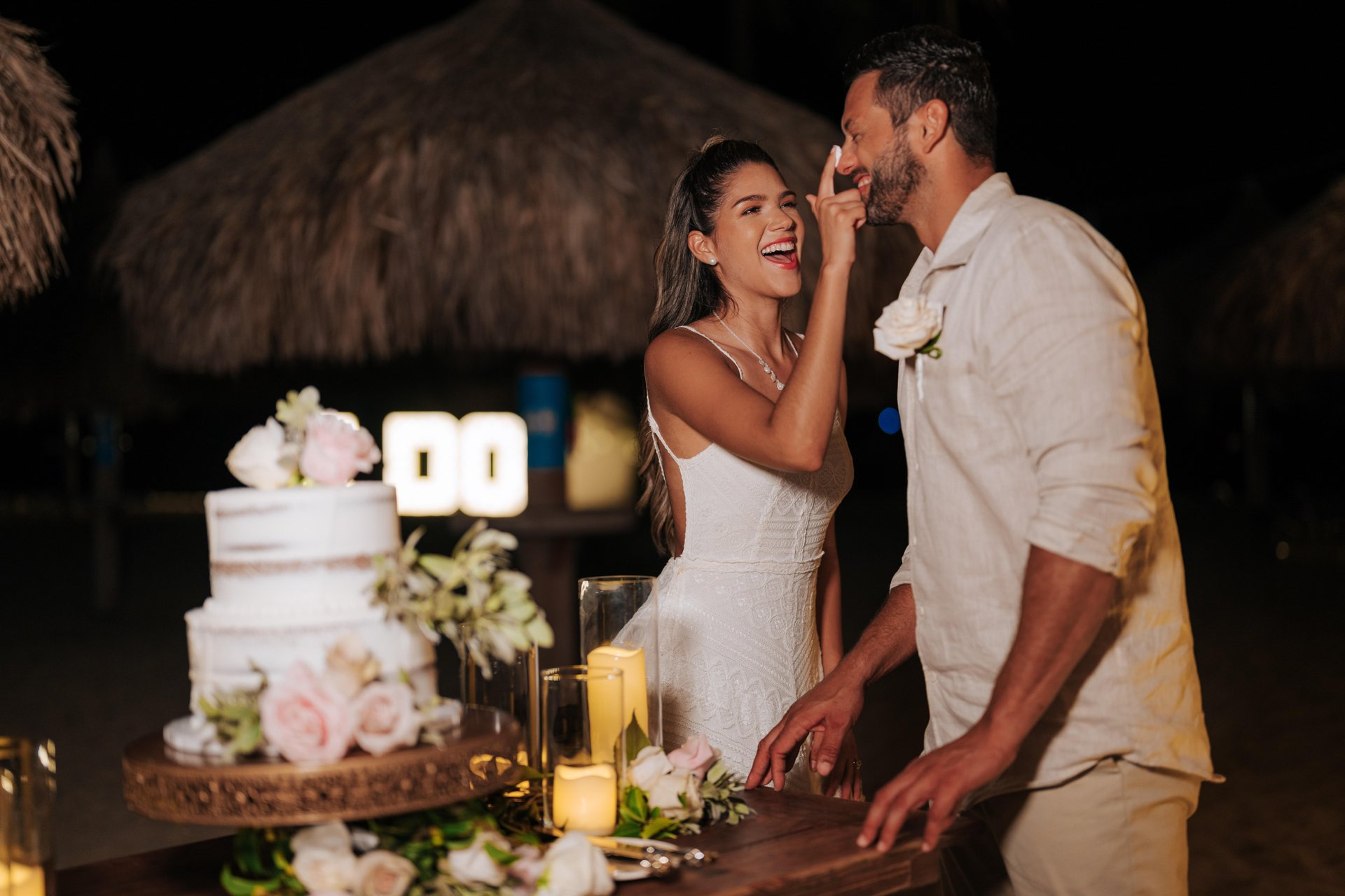 A bride playfully feeds cake to the groom at an outdoor nighttime wedding reception. They are both smiling, standing by a decorated cake table with candles and flowers. Tiki huts are visible in the background.