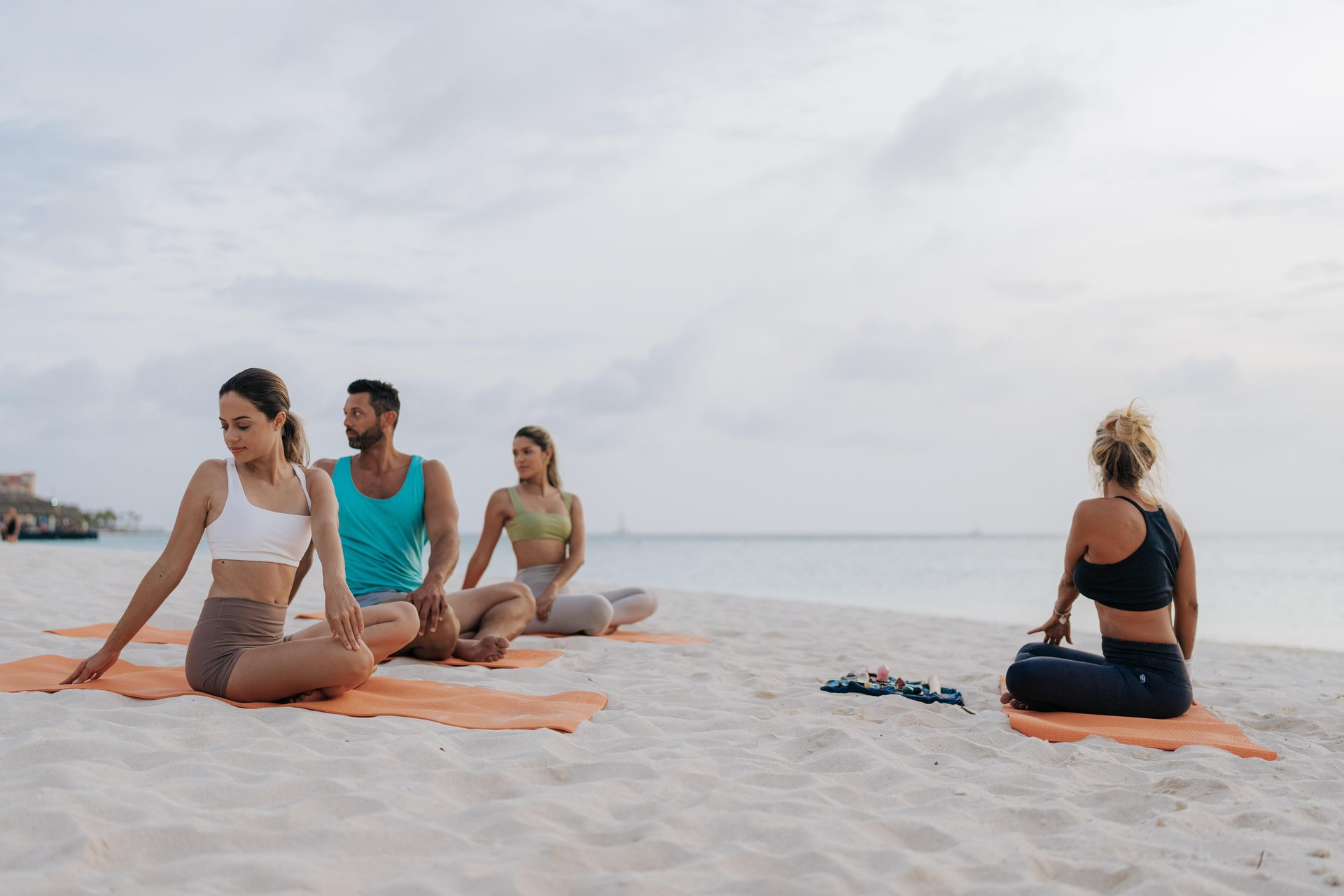 Four people practice yoga on orange mats on a sandy beach near the ocean, performing seated twists. The sky is cloudy, and the atmosphere is calm and peaceful.