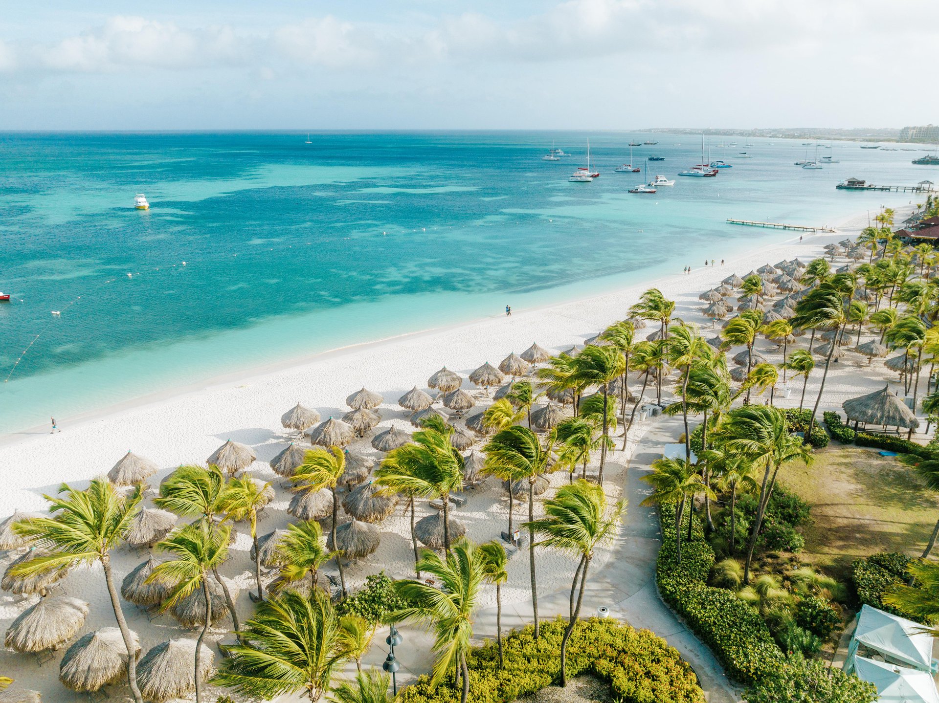 Aerial view of a tropical beach with white sand, turquoise water, palm trees, and thatched umbrellas. Boats are anchored offshore and people are walking along the shoreline under a bright, sunny sky.
