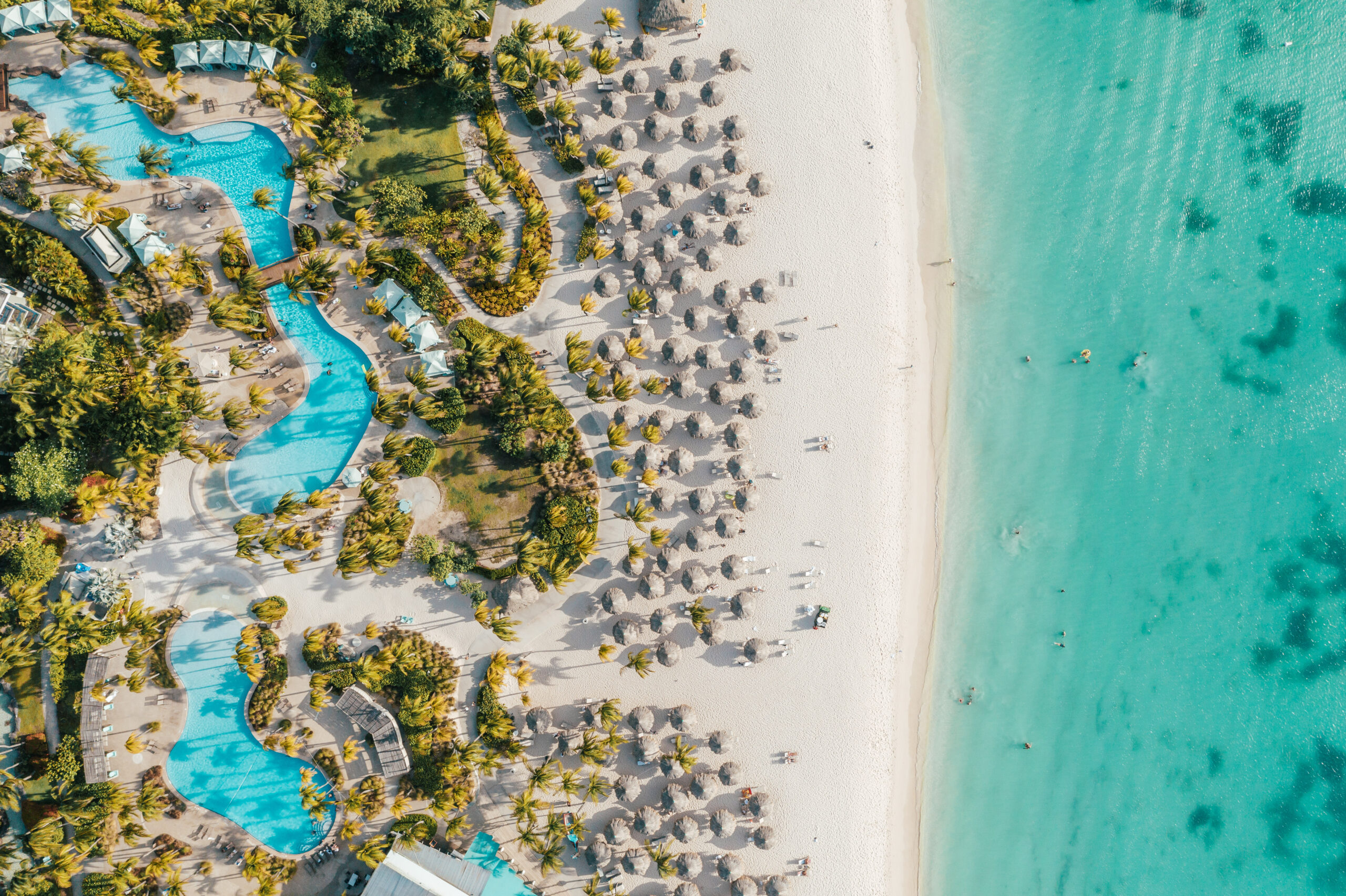 Aerial view of a tropical beach with turquoise water, white sand, palm trees, straw umbrellas, and a winding blue pool surrounded by greenery. People are relaxing along the shoreline and in the water.