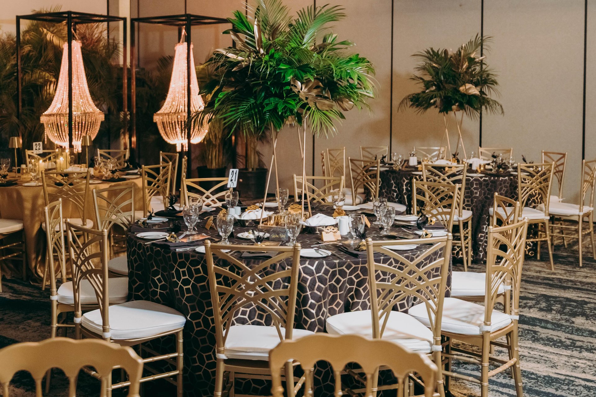 Elegant banquet hall with round tables covered in dark, patterned tablecloths, gold chairs, tall leafy centerpieces, and chandeliers, creating a sophisticated, tropical-inspired event setting.