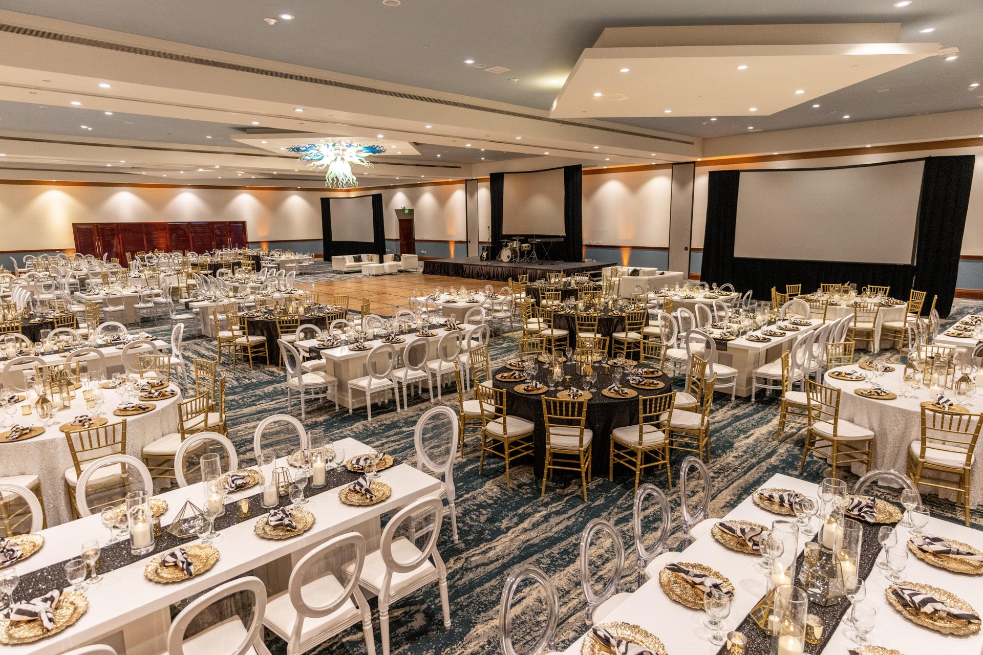 A spacious banquet hall set for an event with round and rectangular tables, elegant place settings, gold and white chairs, candles, and two large projection screens near a dance floor and stage.