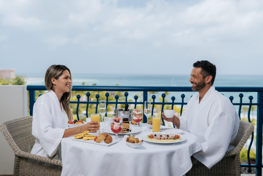 A man and woman in white robes enjoy a breakfast with juice, fruit, and pastries at an outdoor table overlooking the ocean on a sunny day.