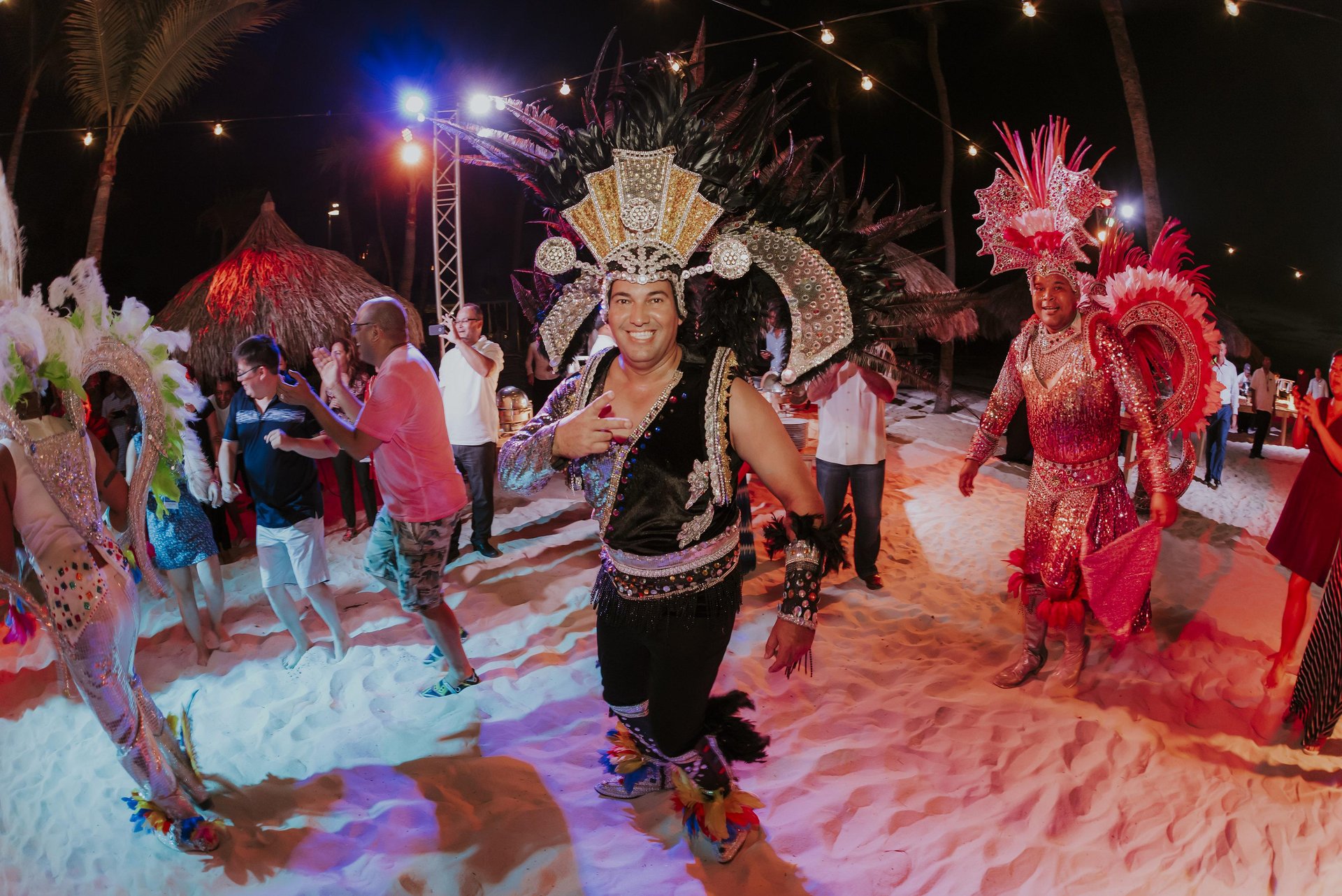 People dance on a sandy beach at night under string lights. The focus is on performers in elaborate, colorful feathered costumes and headdresses, surrounded by other partygoers enjoying the festive atmosphere.