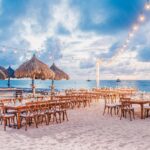 Wooden tables and chairs are set up on a sandy beach under string lights, with straw thatched umbrellas and the ocean in the background. The sky is partly cloudy, and boats are visible on the water.