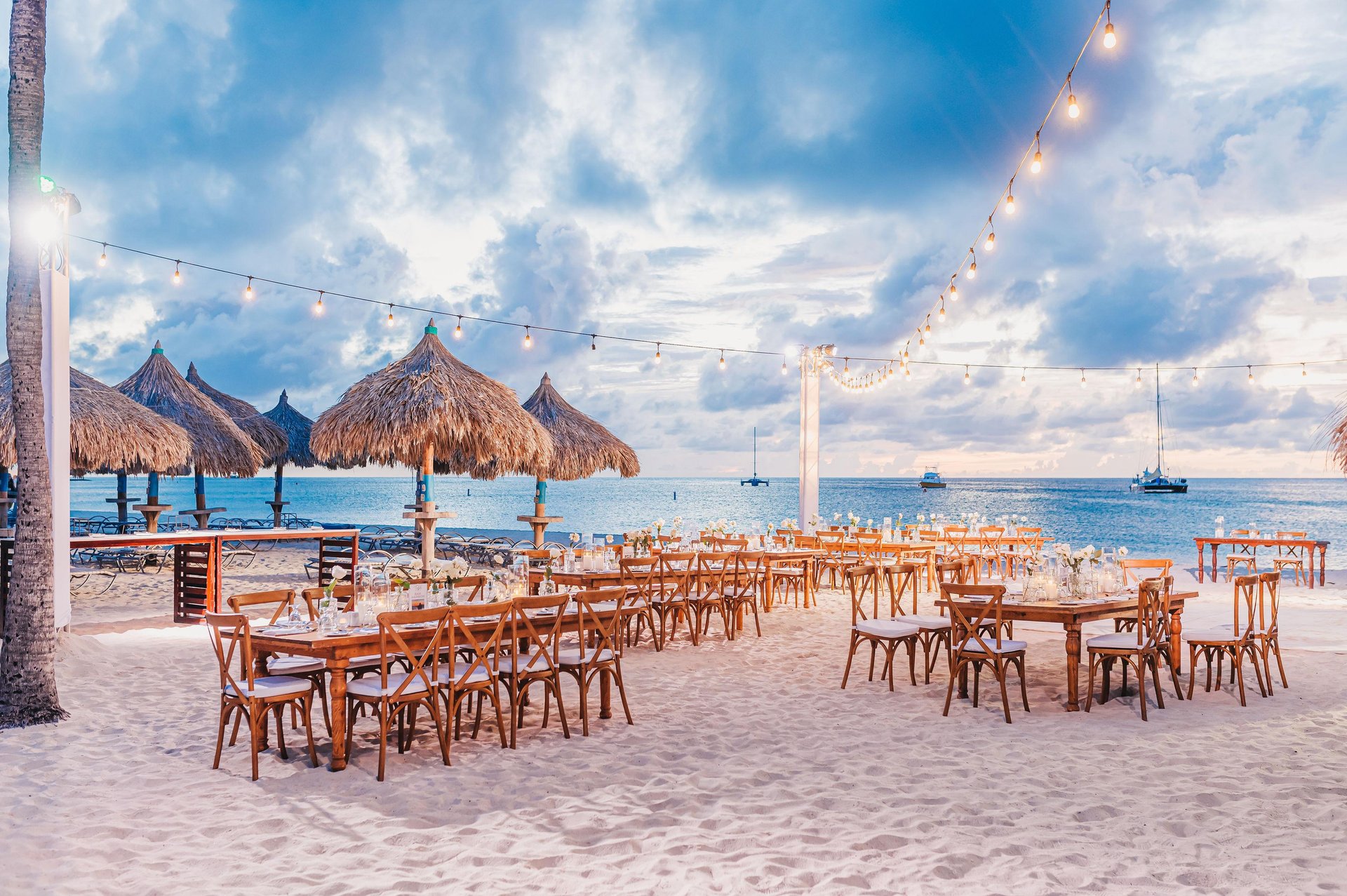 Wooden tables and chairs are set up on a sandy beach under string lights, with straw thatched umbrellas and the ocean in the background. The sky is partly cloudy, and boats are visible on the water.
