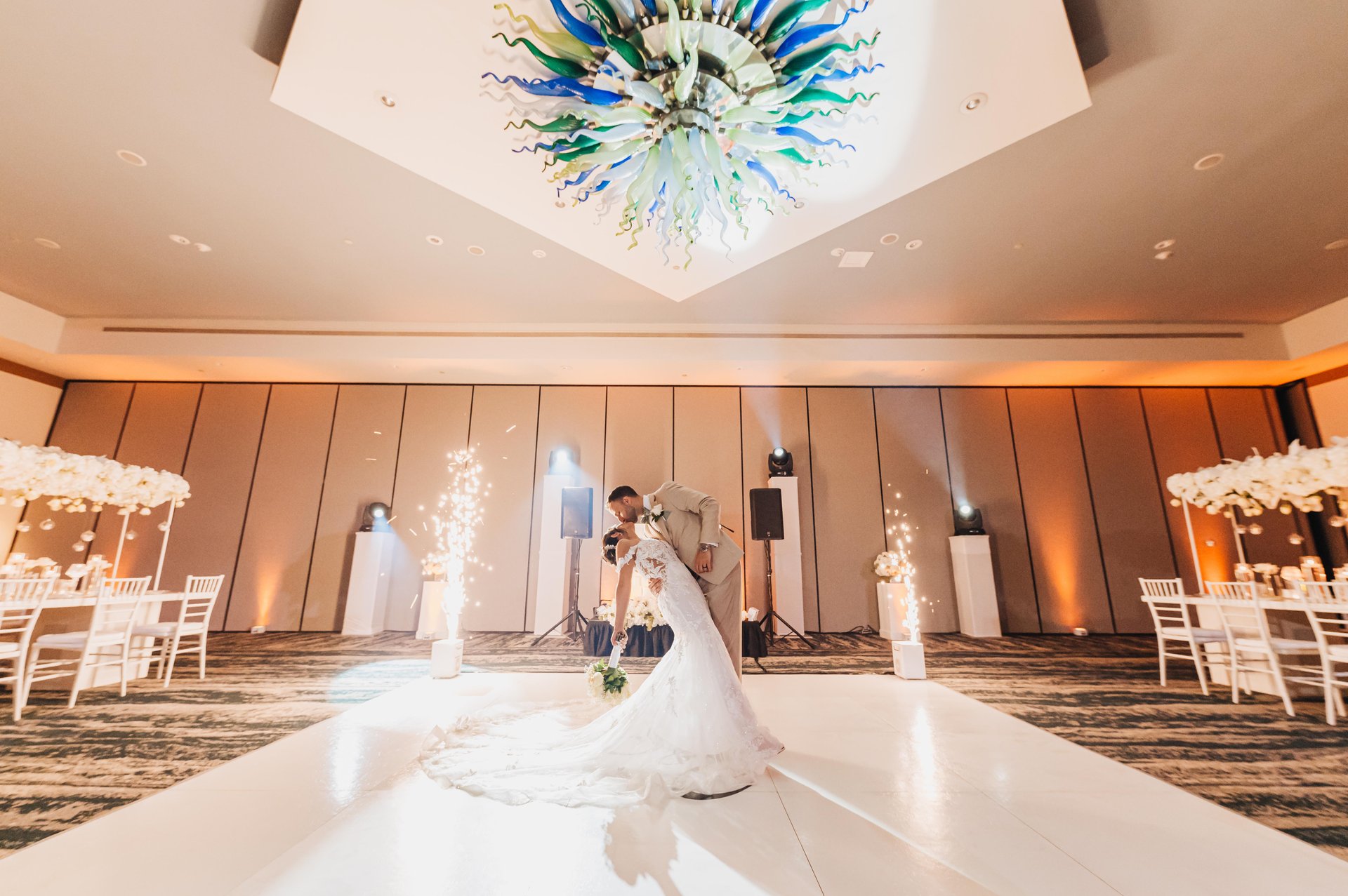 A bride and groom share a romantic dip on a dance floor under a colorful glass chandelier, with white floral decorations and spark fountains illuminating the elegant indoor wedding venue.