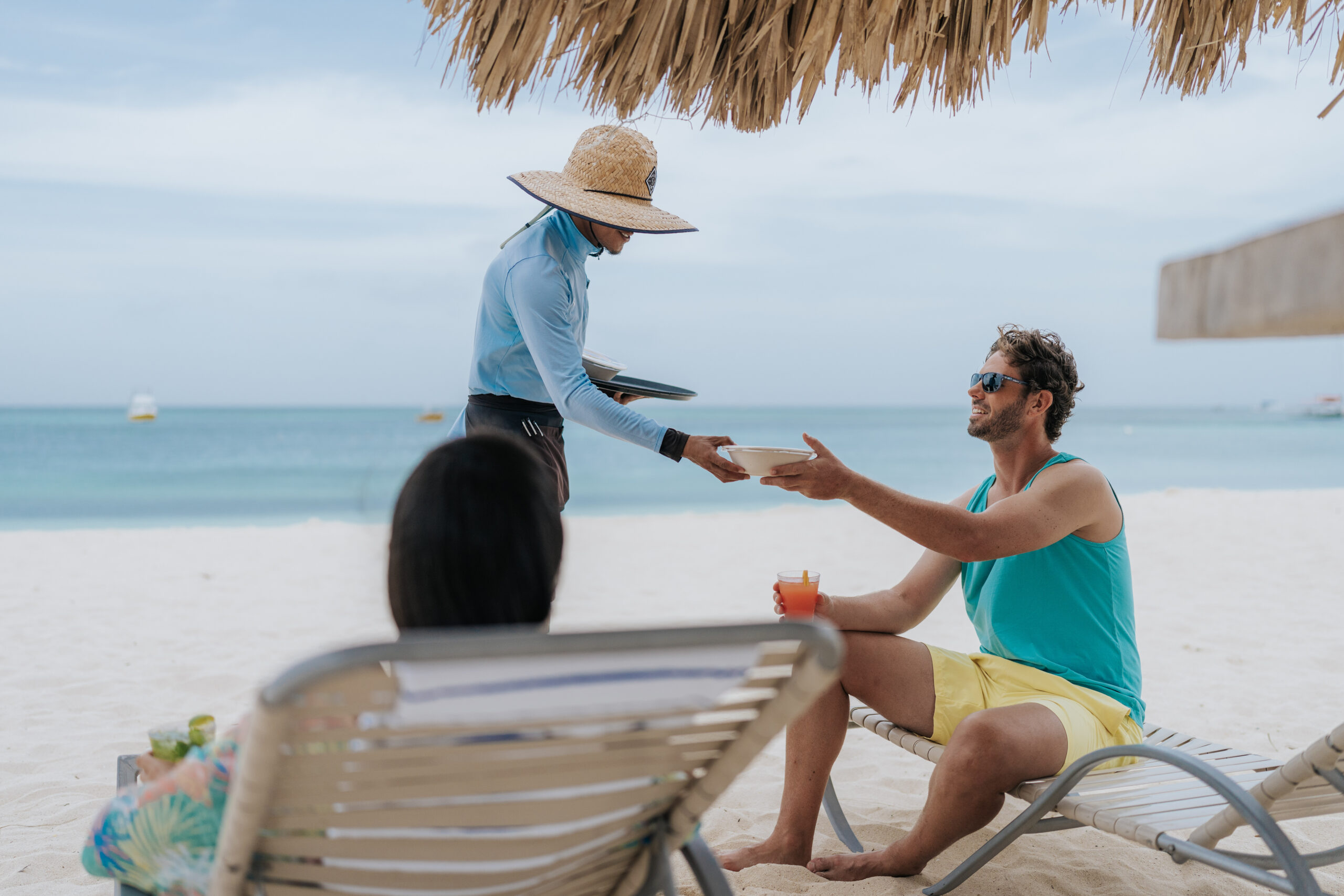 A man in sunglasses and a blue tank top sits on a lounge chair on a sandy beach, receiving a drink from a server wearing a wide hat. Another person is seated nearby, facing the ocean.