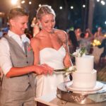 Two brides, one in a white suit vest and the other in a strapless wedding dress, smile as they cut a white tiered cake together at an outdoor evening wedding reception. Guests and string lights are in the background.