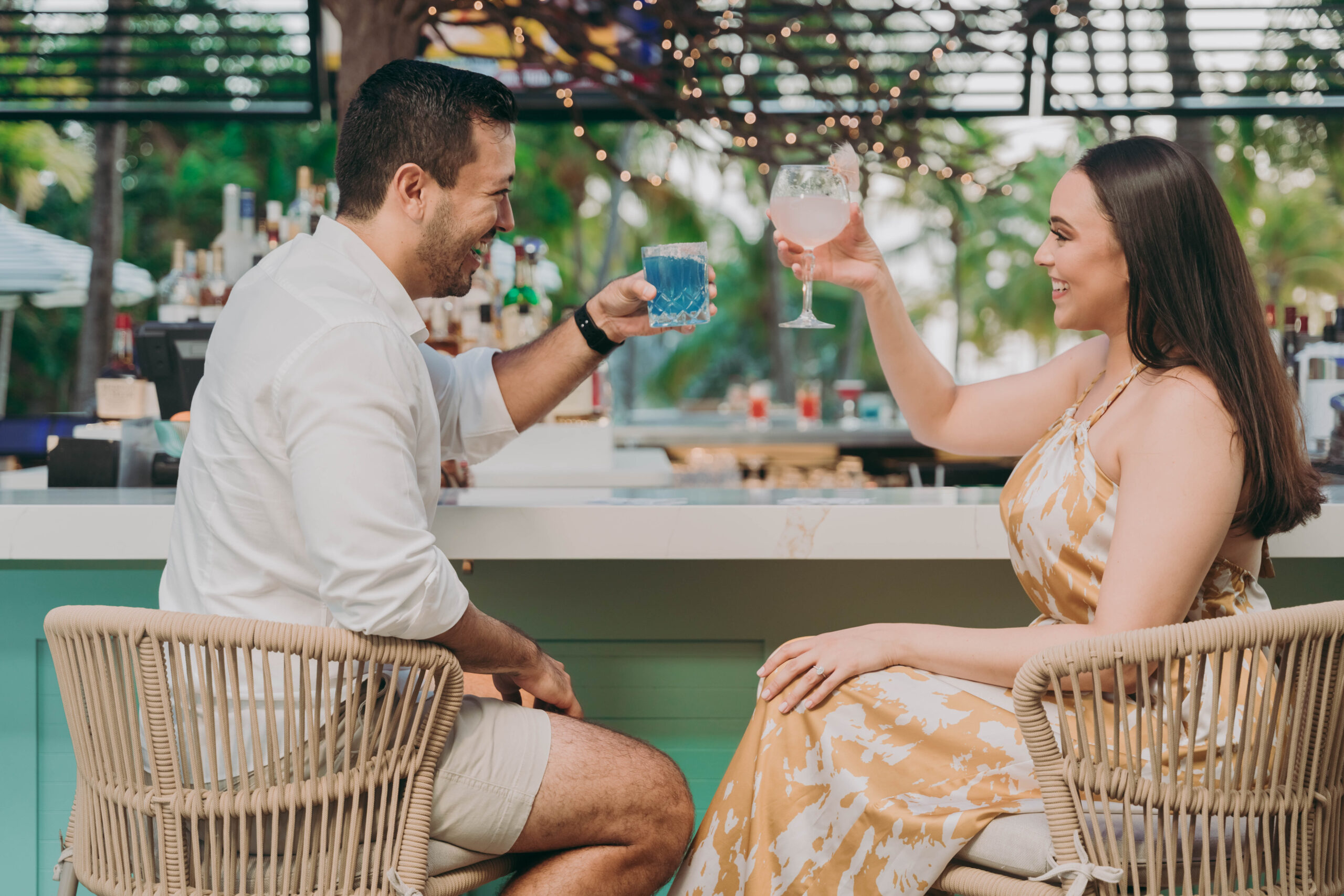 A man and a woman sit at an outdoor bar, smiling and toasting with colorful drinks. The atmosphere is relaxed, with string lights and greenery in the background.