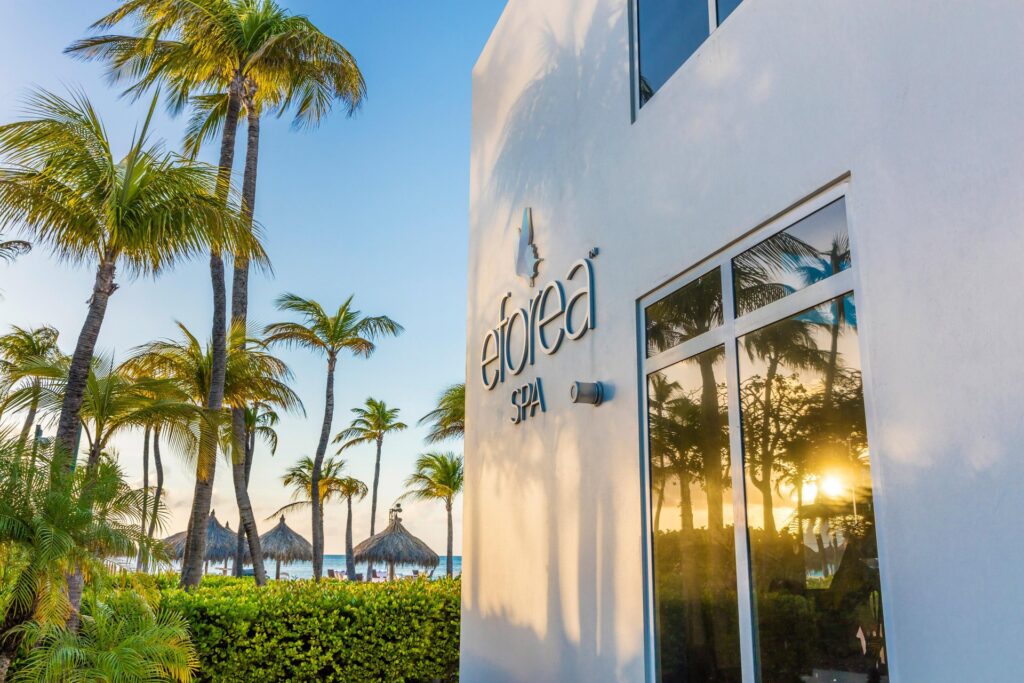 A modern white building with a sign reading “eforea spa” is surrounded by palm trees. Thatched beach umbrellas and the ocean are visible in the background under a clear blue sky. The spa’s window reflects the scenic view.