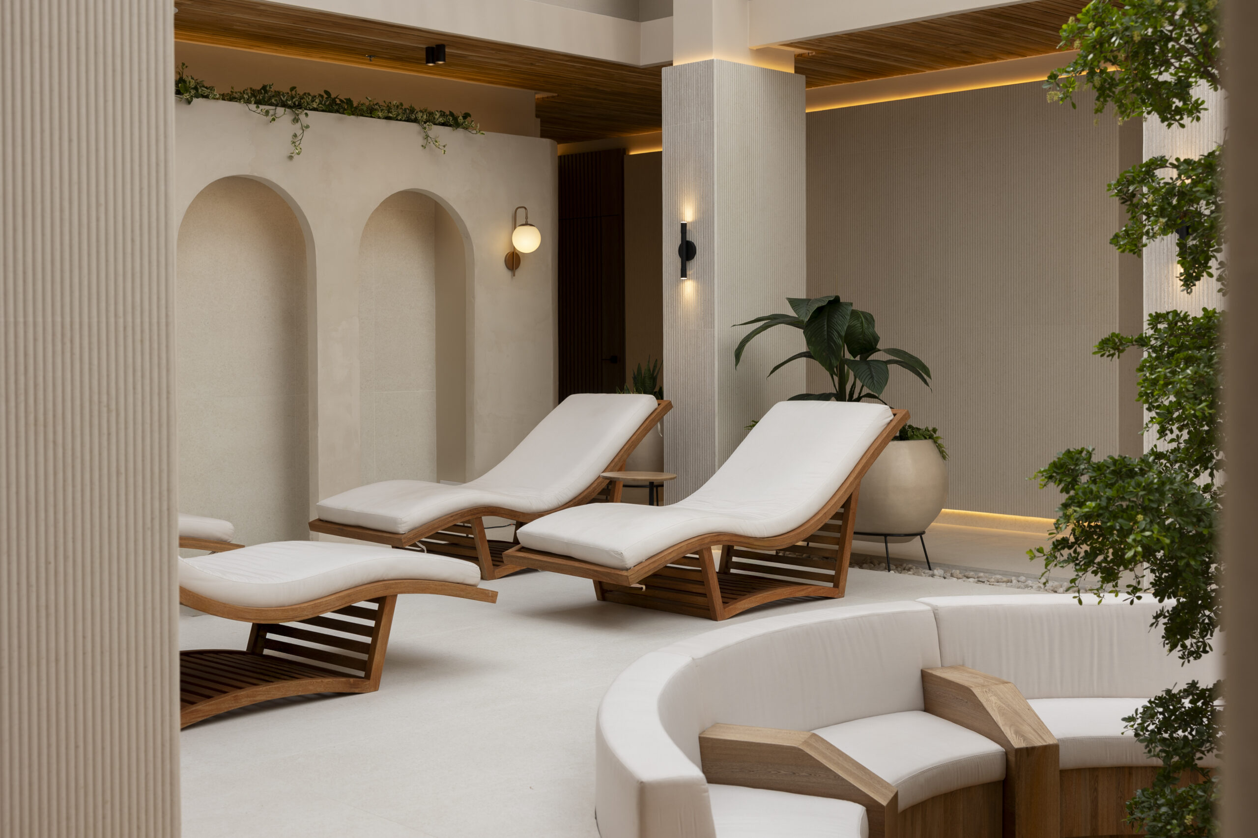 Two modern wooden lounge chairs with white cushions sit in a serene, minimalist indoor space with arched wall niches, potted plants, and warm ambient lighting, creating a tranquil, spa-like atmosphere.