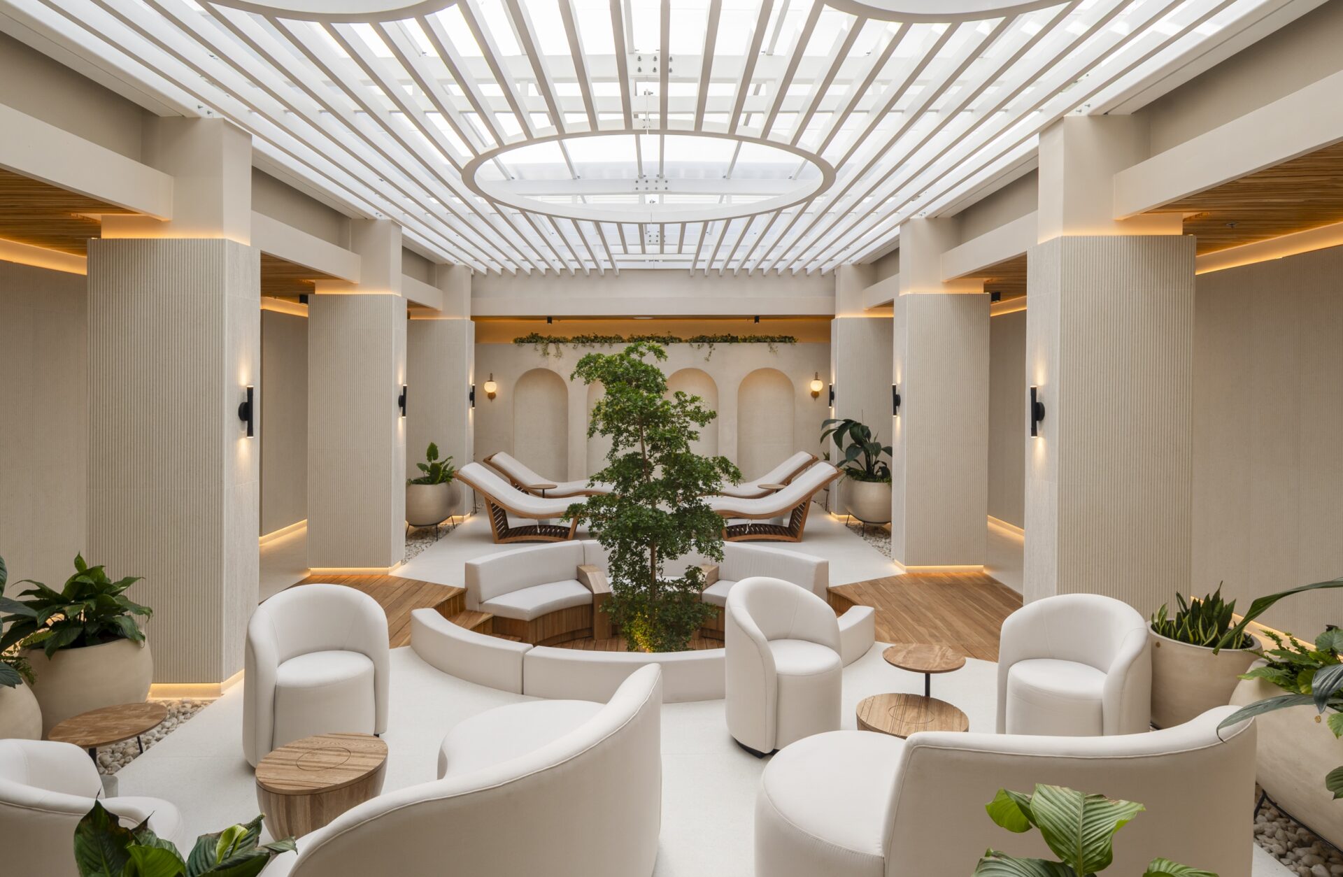 A modern, spacious lounge with white chairs, circular sofas, and wooden tables. Sunlight streams through a large skylight. Green plants and trees decorate the room, creating a calm and inviting atmosphere.