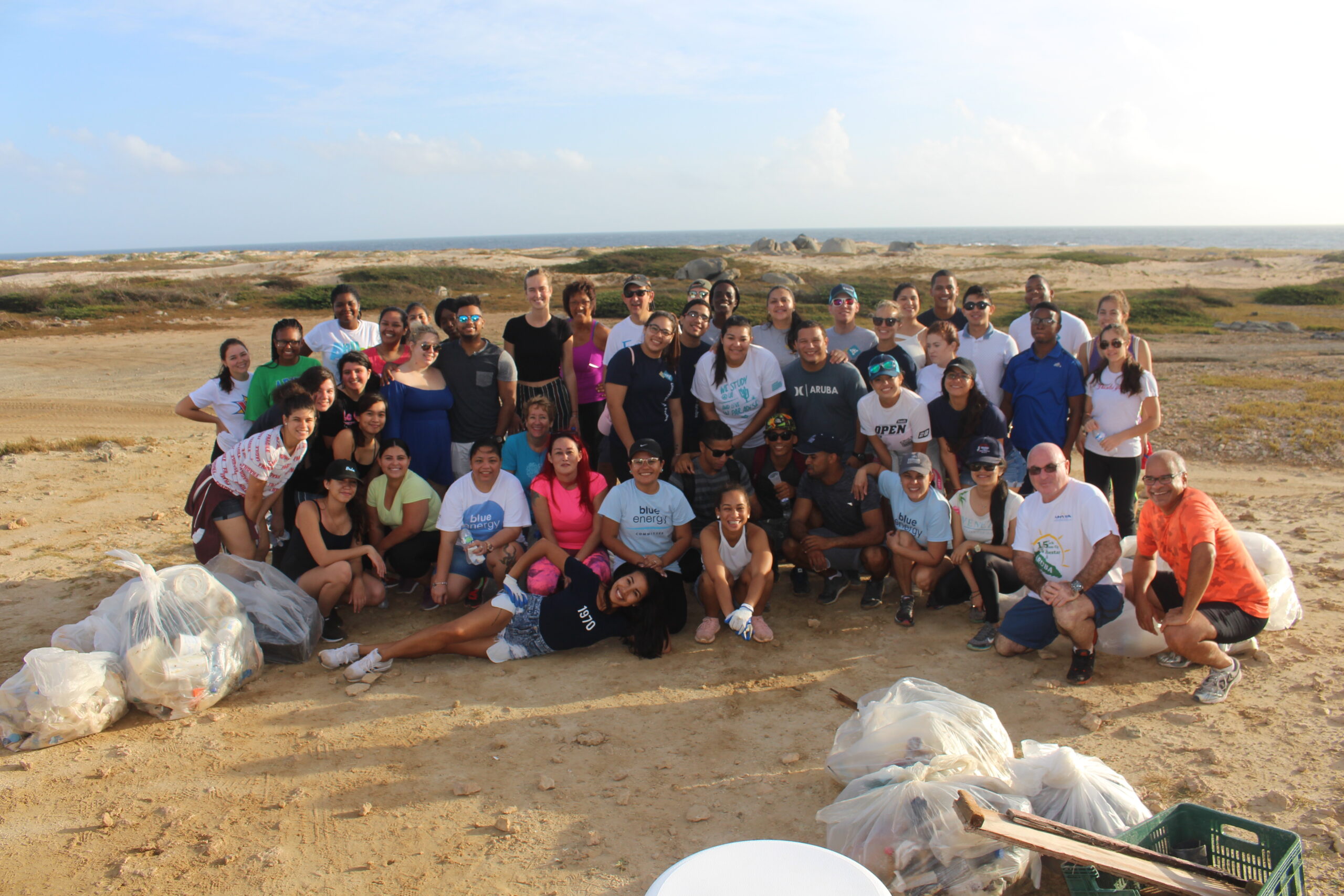 A large group of people of various ages pose and smile on a sandy beach with several bags of collected trash, suggesting a beach clean-up event. The ocean and blue sky are visible in the background.