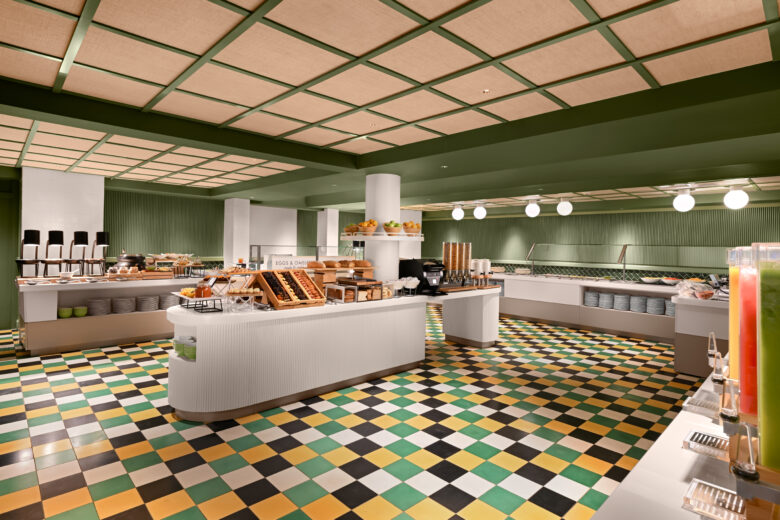 A brightly lit buffet restaurant features colorful checkered floor tiles, white display counters with assorted pastries, fruit, juice dispensers, and cereal stations, with neatly stacked plates along the back wall.