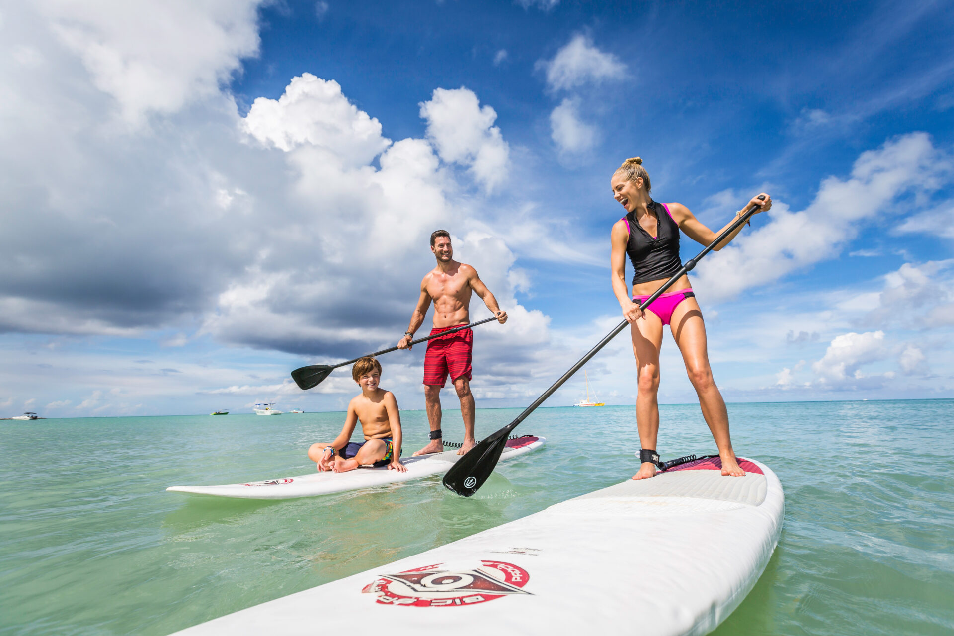 A woman, a man, and a child paddleboarding on a calm, turquoise sea under a blue sky with scattered clouds. The woman and man are standing, while the child kneels on their board, all enjoying the water.
