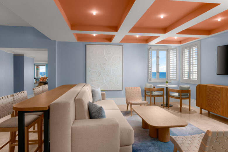 A modern living room with light blue walls, an orange coffered ceiling, a beige sofa, wooden chairs and tables, abstract wall art, and large windows with white shutters overlooking the ocean.