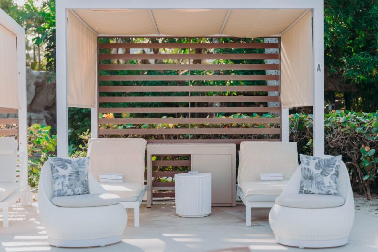 Poolside cabana with light-colored lounge chairs, round chairs with leaf-patterned pillows, a small round table, and wooden slat backdrop, surrounded by lush greenery in bright sunlight.