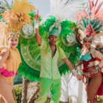 Three people in vibrant, colorful festival costumes with large feathered headdresses and wings pose and smile outdoors, surrounded by palm trees and bright sunlight.