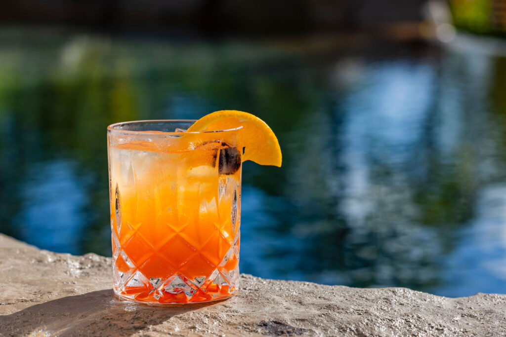 A colorful cocktail with ice, garnished with an orange slice and cherry, sits on a stone ledge beside a blurred blue swimming pool in bright sunlight.