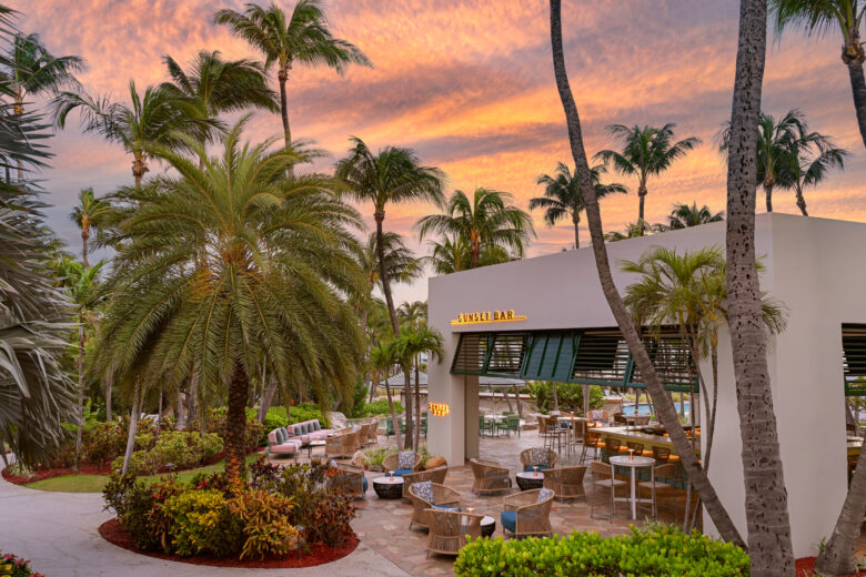 Outdoor lounge area with wicker chairs and tables under palm trees at sunset, beside a modern bar building with a yellow "SUNSET BAR" sign, lush greenery, and a colorful sky in the background.