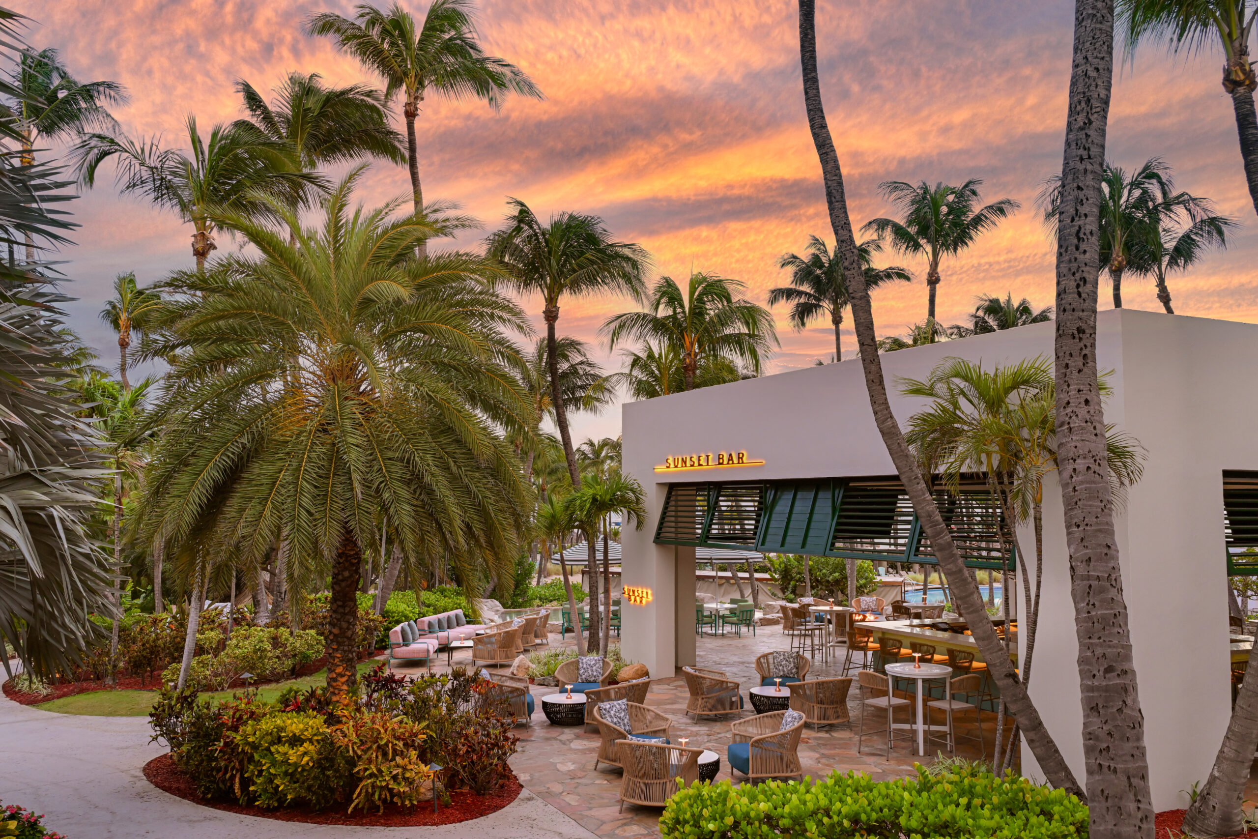 Outdoor lounge area with wicker chairs and tables under palm trees at sunset, beside a modern bar building with a yellow "SUNSET BAR" sign, lush greenery, and a colorful sky in the background.